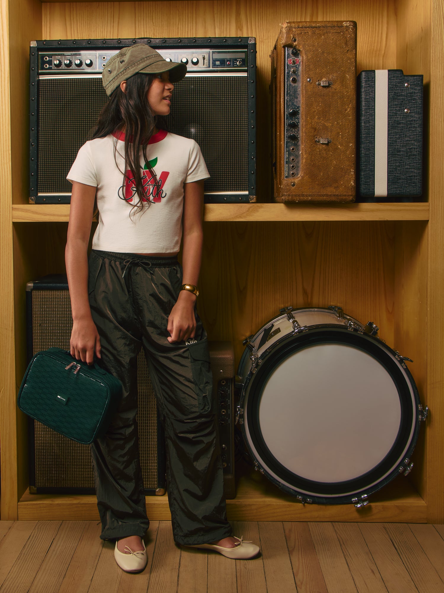 Kith Kids girl in Kith NY apple ringer tee and black cargo pants holds a green Kith monogram pouch among vintage amps
