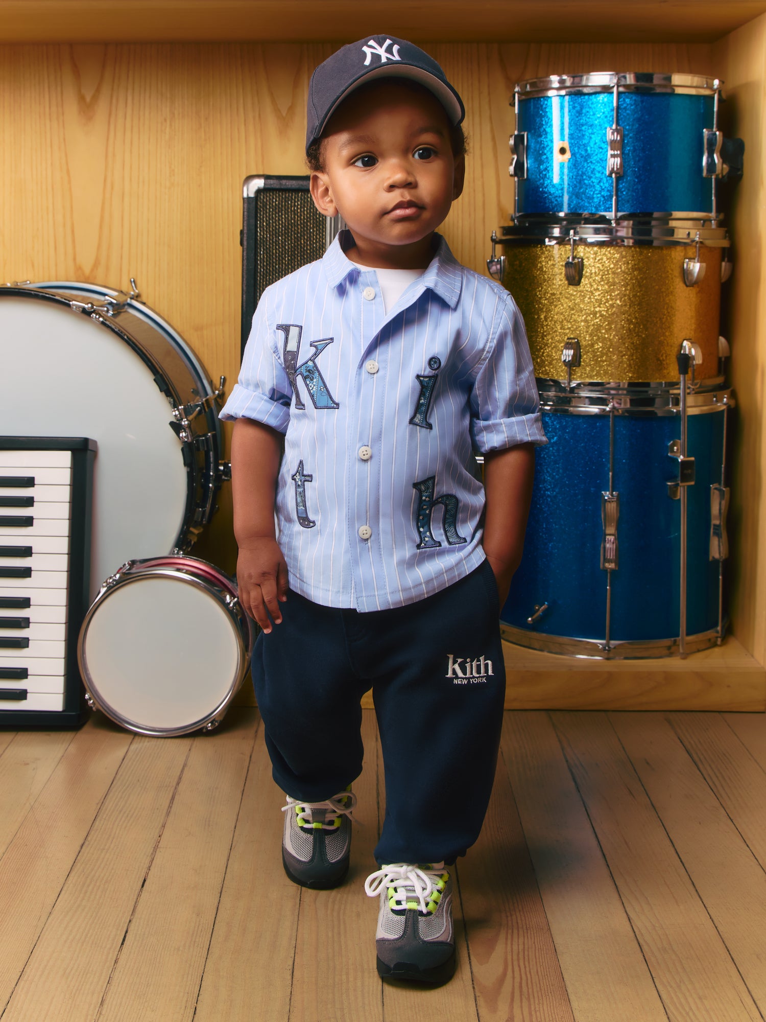 Kith Kids toddler boy in Yankees cap, kith-appliqué pinstripe shirt and navy Kith sweatpants by a glitter drum kit