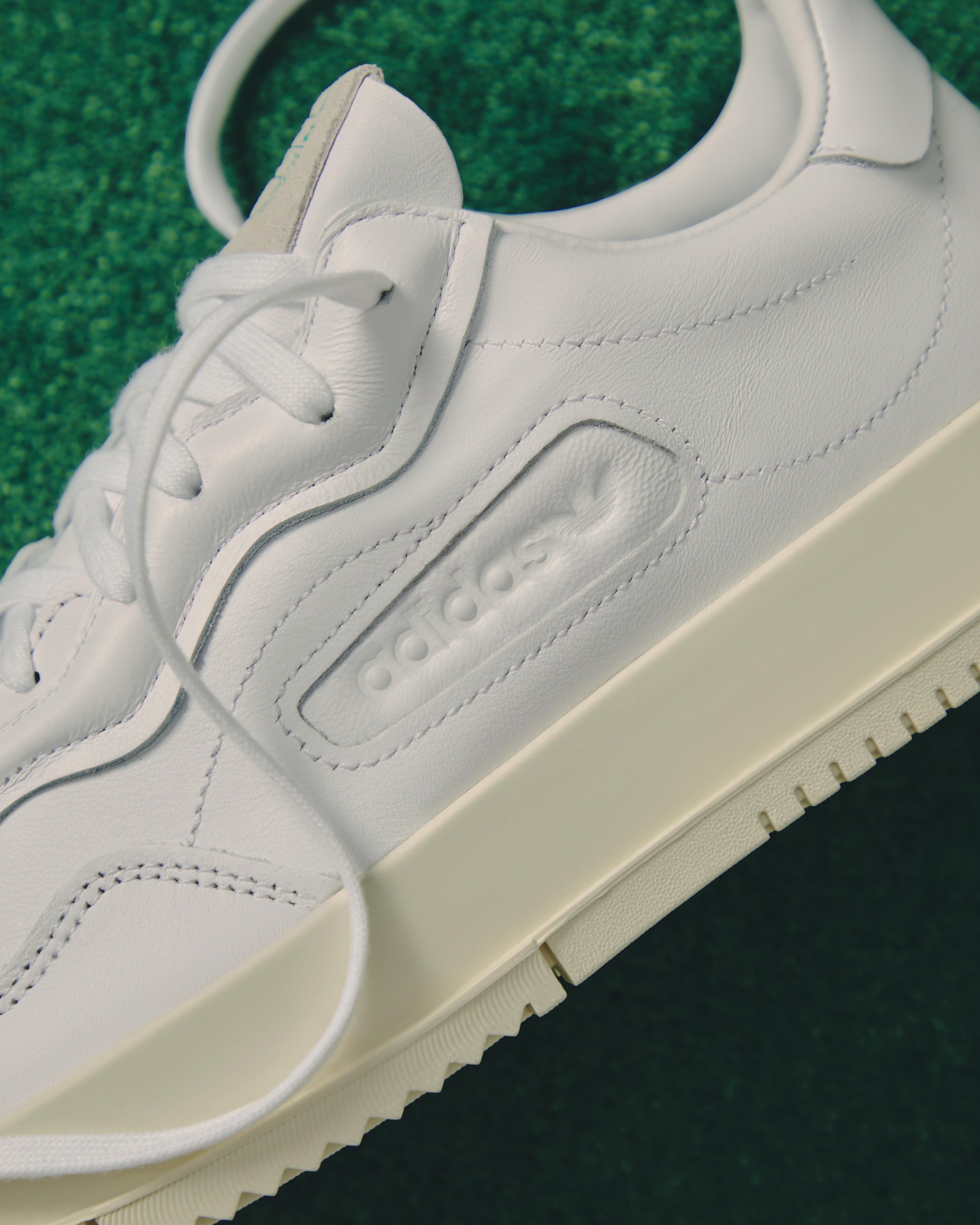 Close-up of a white leather adidas SC Premiere sneaker with embossed logo and trefoil, resting on green turf