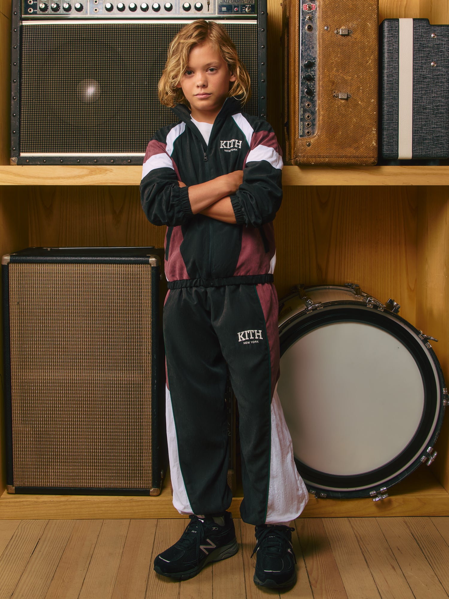 Kith Kids boy, arms crossed, in burgundy and black colorblock Kith New York velour tracksuit among vintage amps