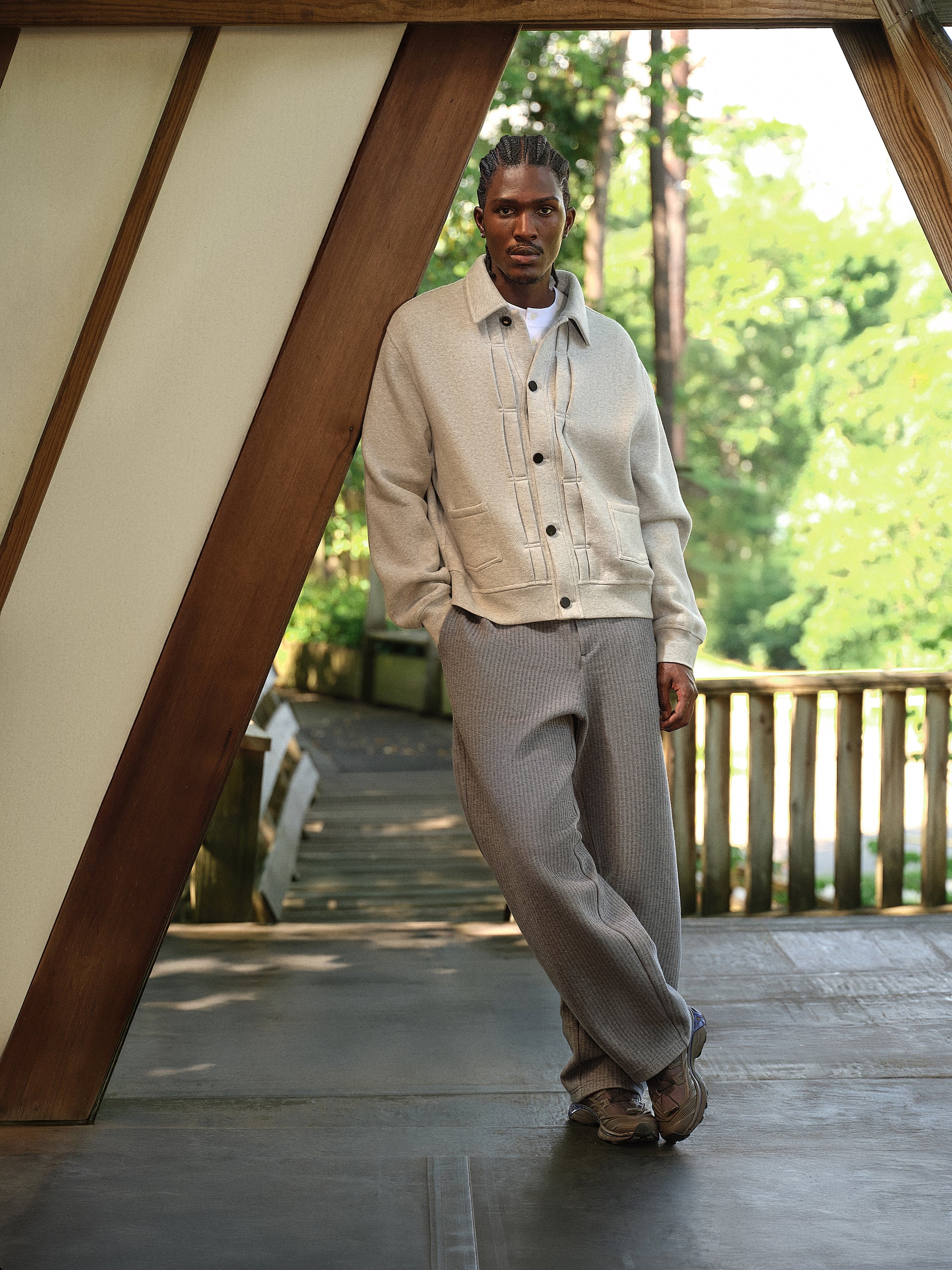 Model in a cream Kith knit overshirt and grey ribbed trousers standing on a wooden footbridge among green trees