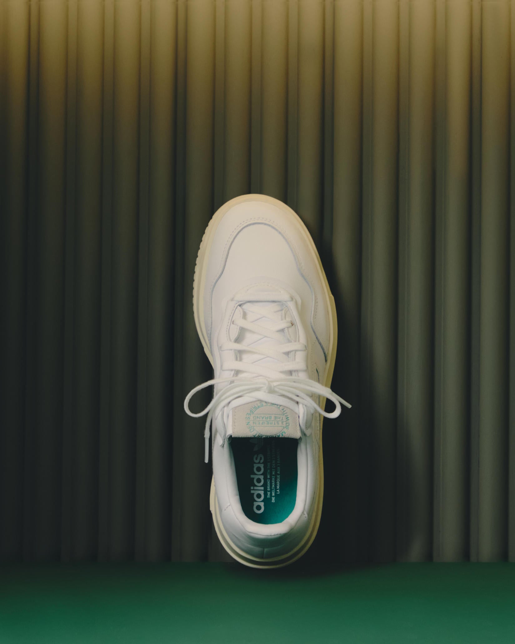 Top-down view of a single white leather adidas SC Premiere sneaker with green insole against a fluted green backdrop