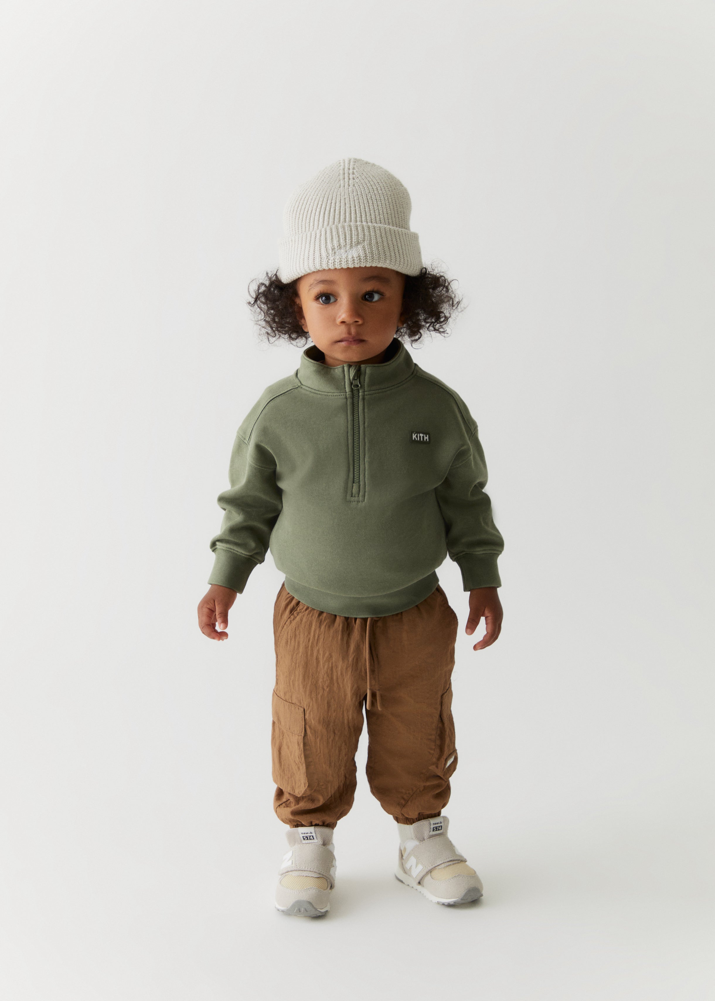 A toddler wearing a Kith script knit beanie, a green quarter-zip sweater, and brown cargo pants from the Kith Kids Fall Classics 2023 collection.