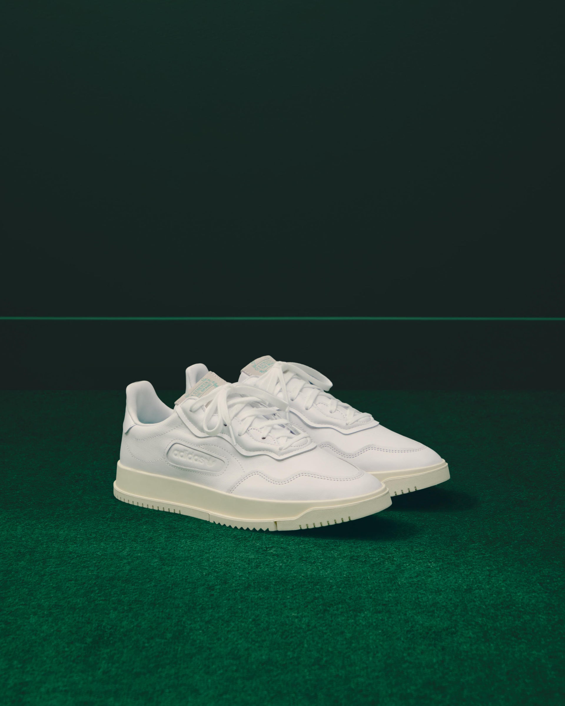 Pair of white leather adidas SC Premiere sneakers with tonal three-stripes on green turf against a dark green backdrop