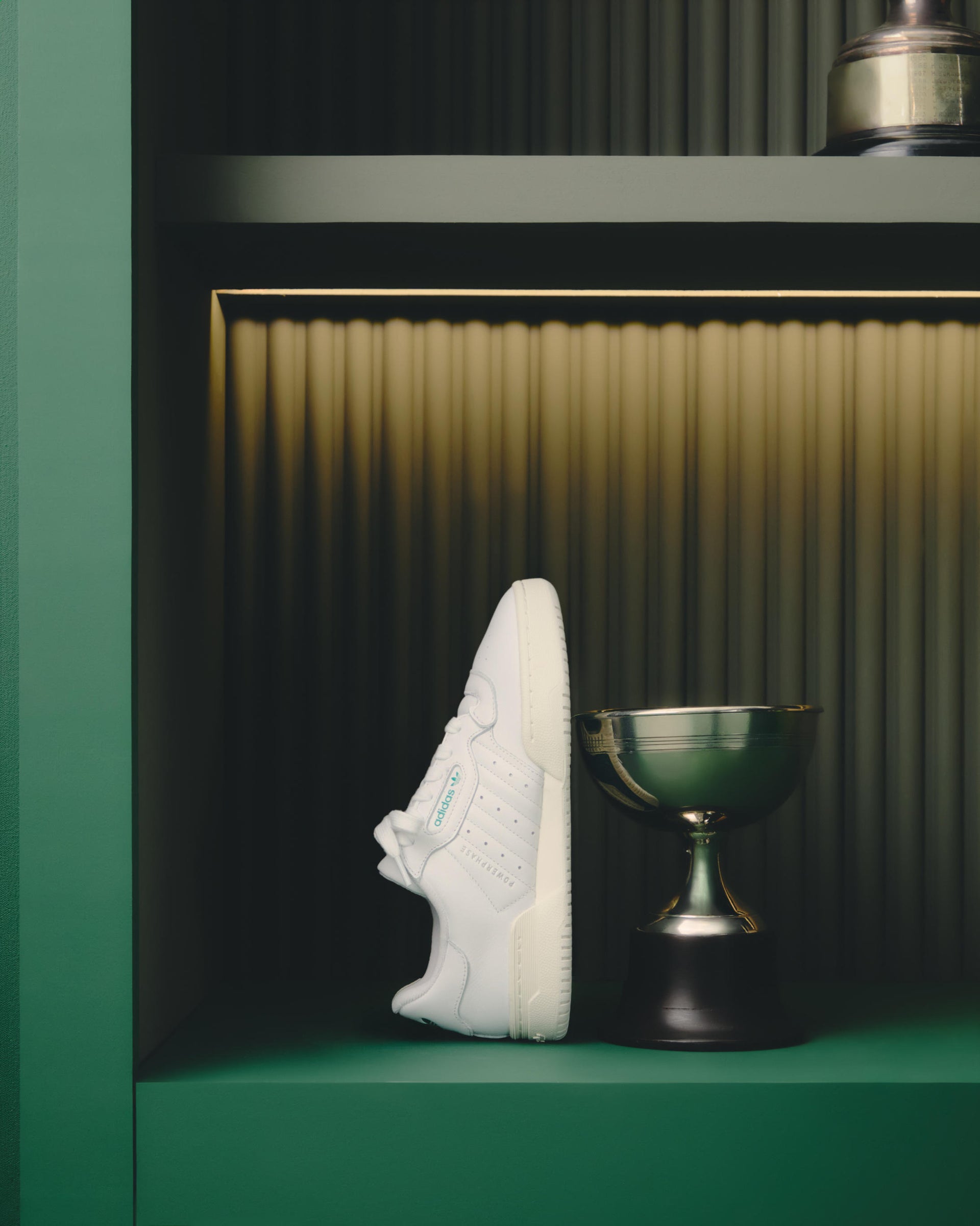 Single white adidas Powerphase sneaker with green trim displayed on a green shelf niche beside a brass trophy chalice