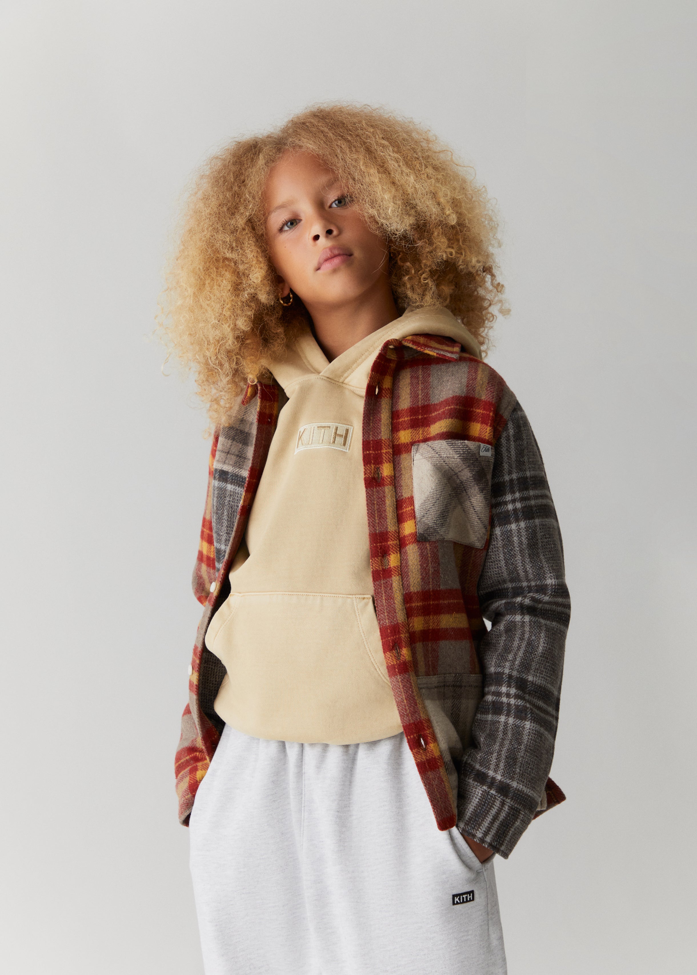A child wearing a color-blocked flannel, Kith-branded tan hoodie, and Kith-branded grey sweatpants from the Kith Kids Fall Classics 2023 collection.