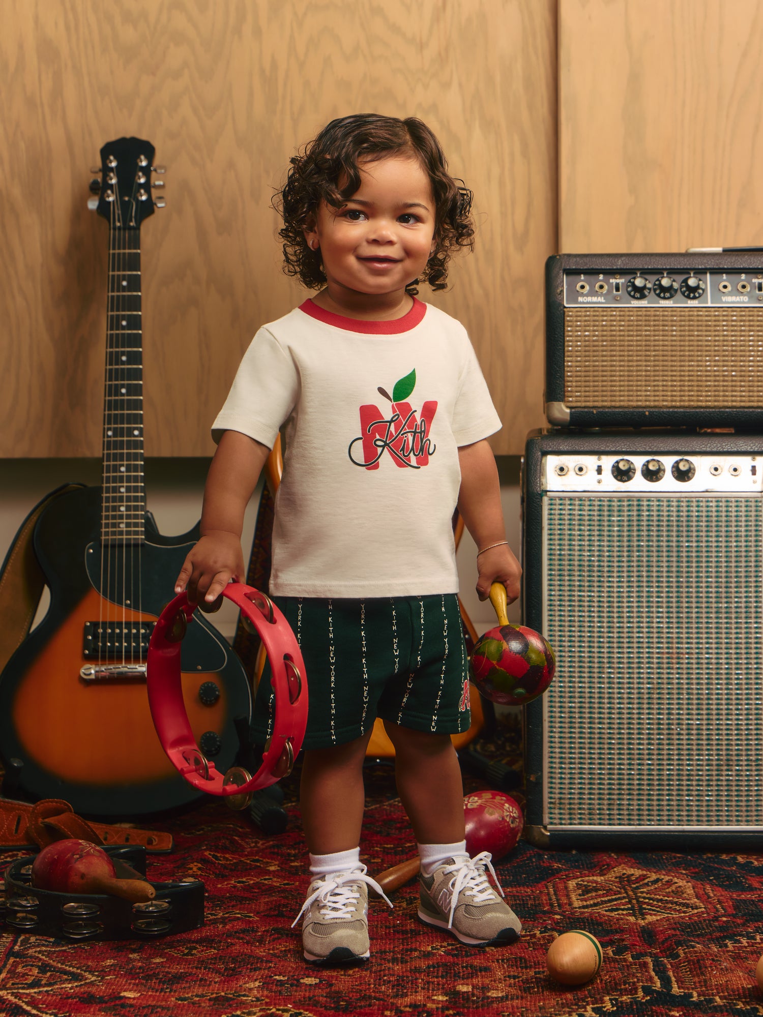 Kith Kids toddler in Kith NY apple ringer tee and green Kith shorts holds a tambourine and maraca among guitars and amps