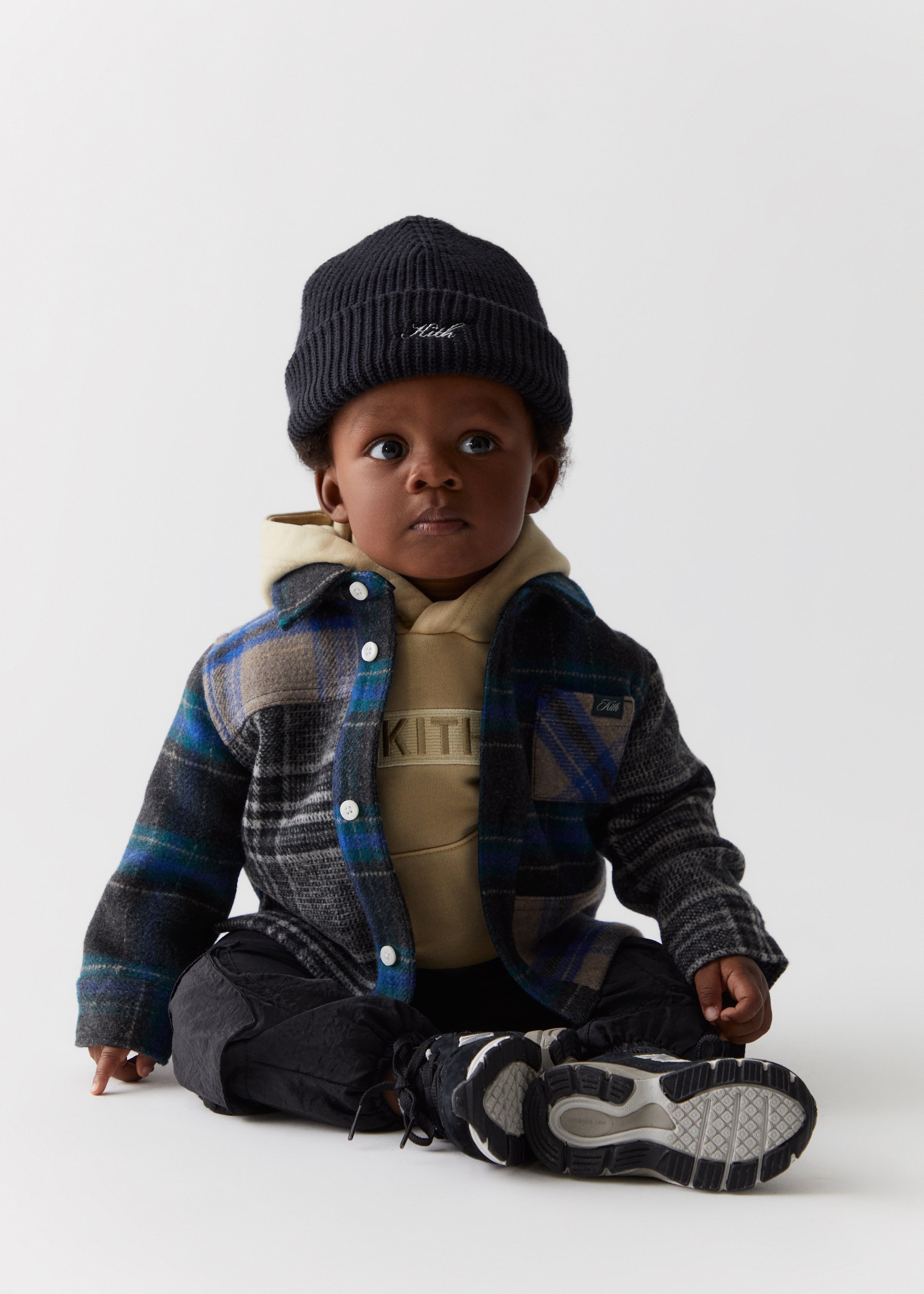 A baby wearing a black knit beanie, a blue flannel overshirt, tan Kith-branded hoodie, and black pants from the Kith Kids Fall Classics 2023 collection.