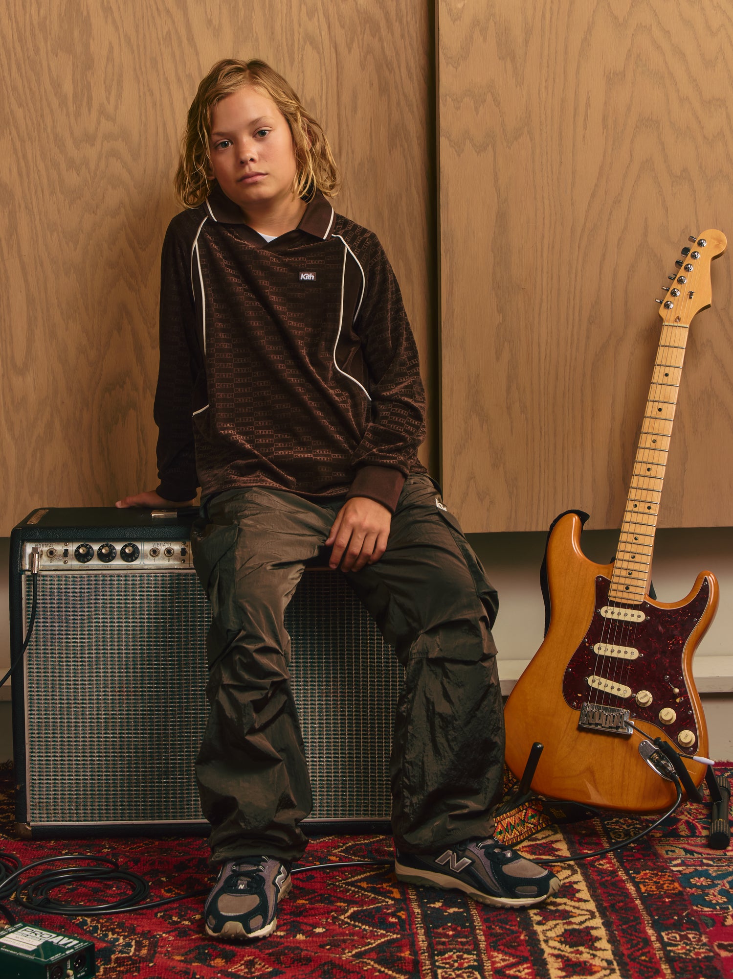 Kith Kids blond boy on a vintage amp in brown Kith monogram rugby and olive cargo pants beside a Fender Stratocaster