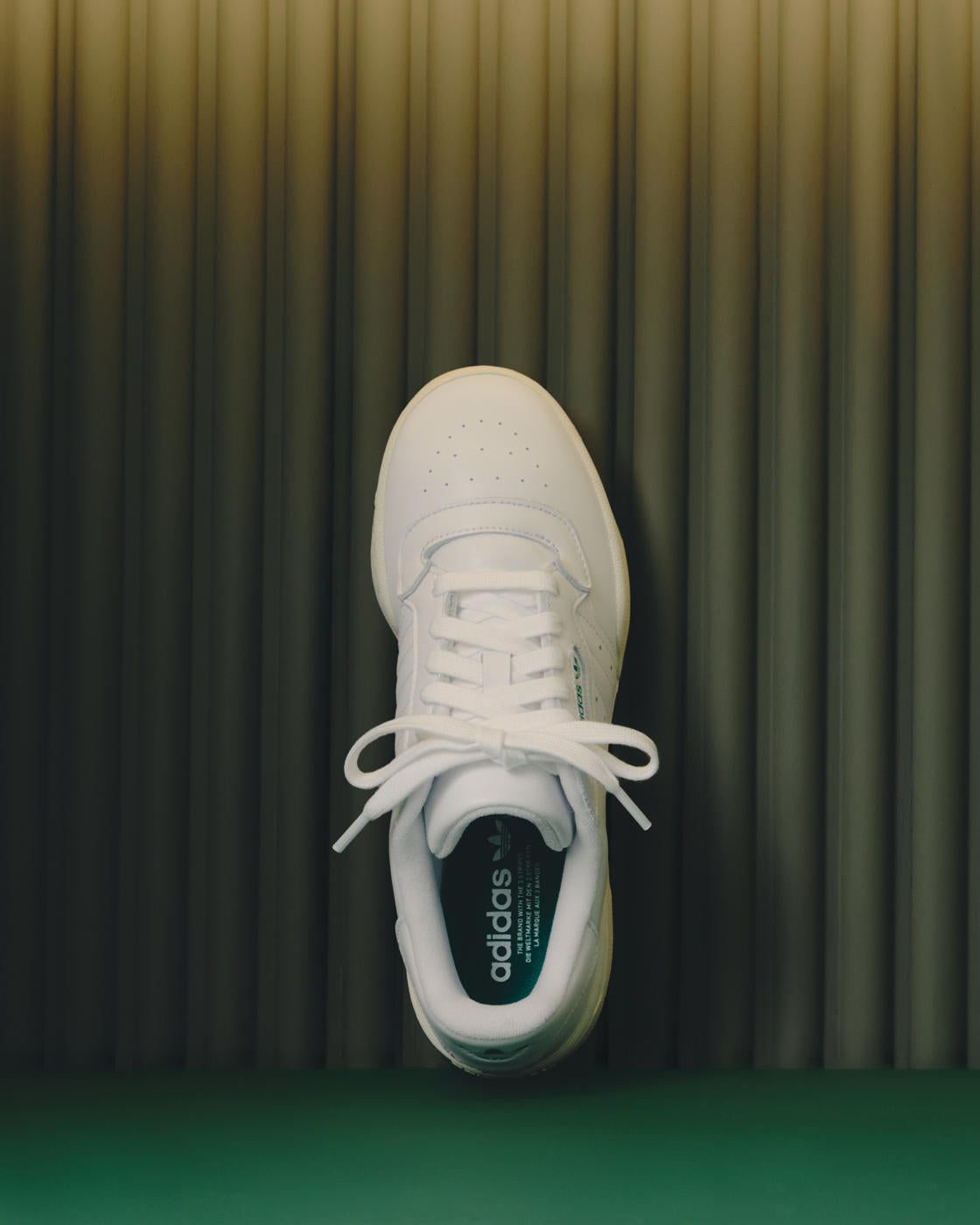 Top-down view of a single white adidas leather sneaker with green heel accents against a ribbed olive curtain