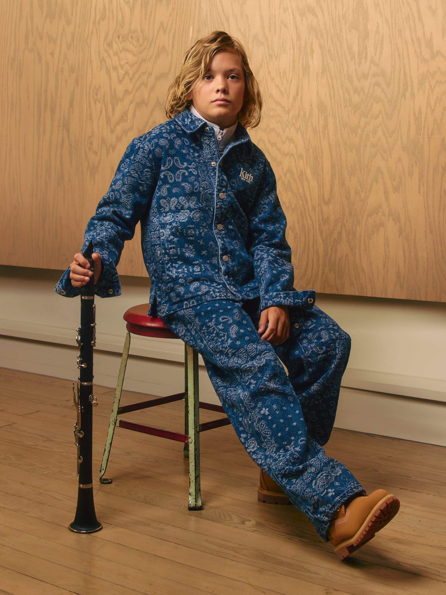 Blond boy on a stool in a blue paisley Kith denim jacket and matching pants, holding a clarinet, tan boots against a wood-panel backdrop
