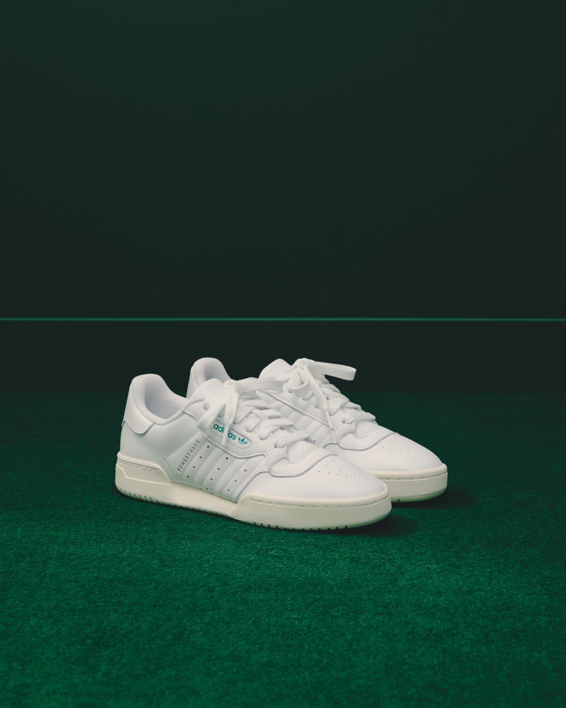 Pair of white adidas Powerphase leather sneakers with green accents, front angle, on green carpet with a dark backdrop