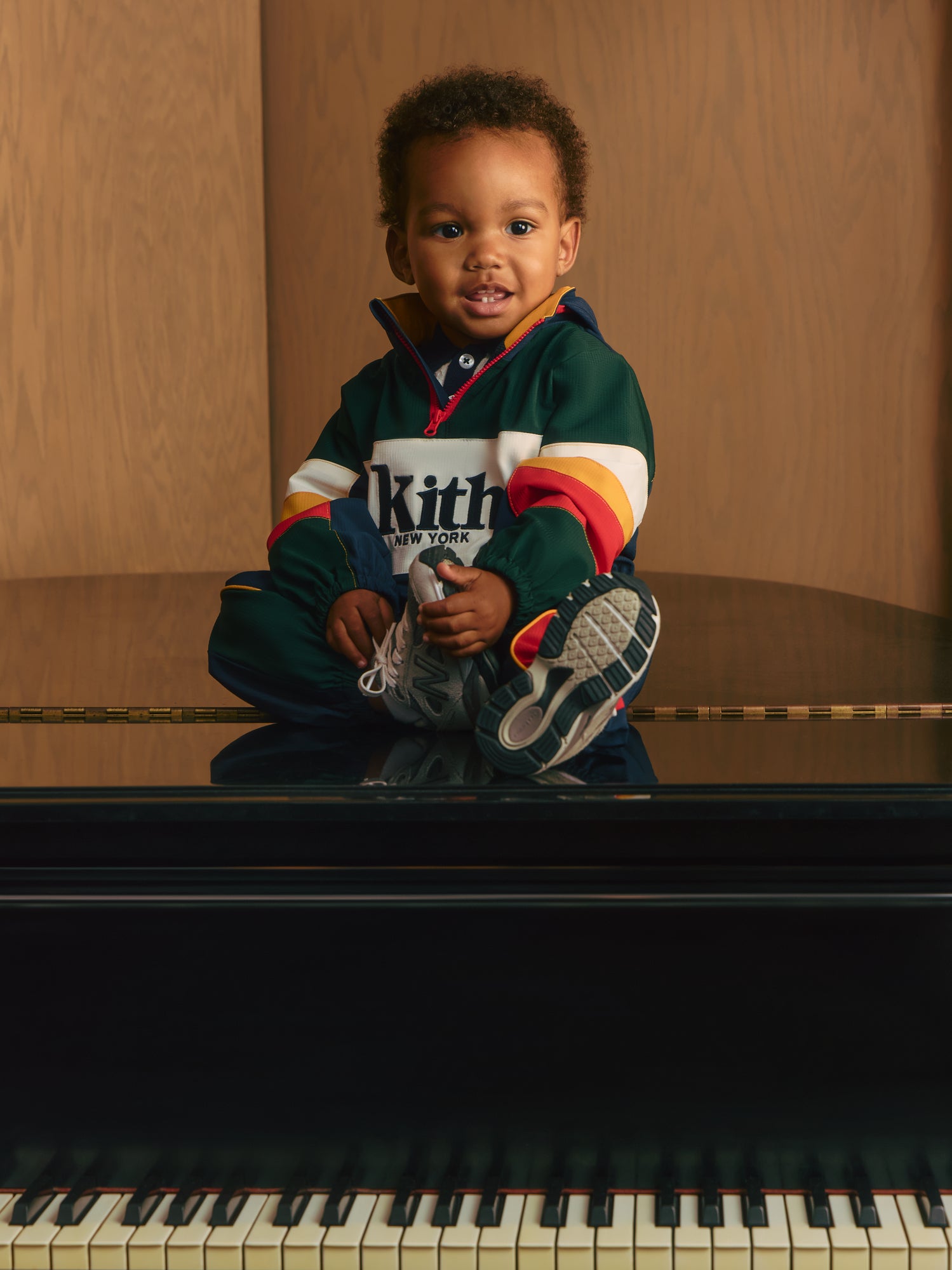 Smiling toddler boy seated atop a black grand piano in a green, red and cream Kith New York colorblock zip pullover and New Balance sneakers