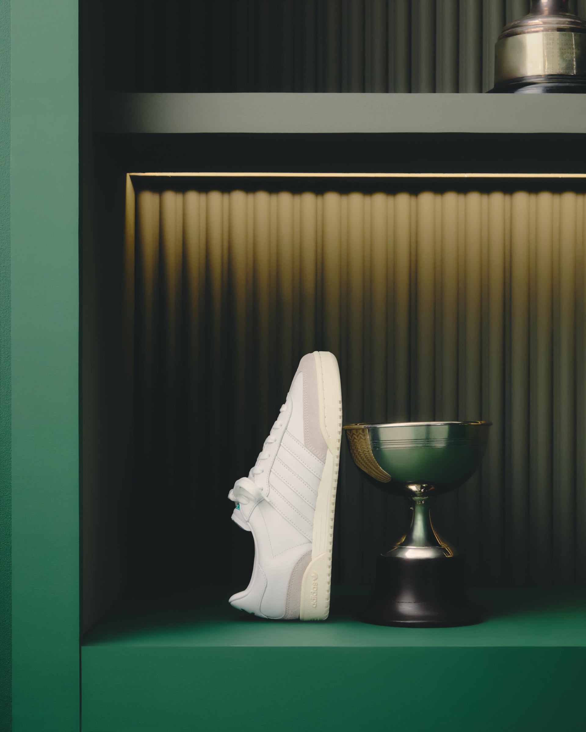 White adidas leather sneaker in side profile displayed on a green shelf beside a gold trophy