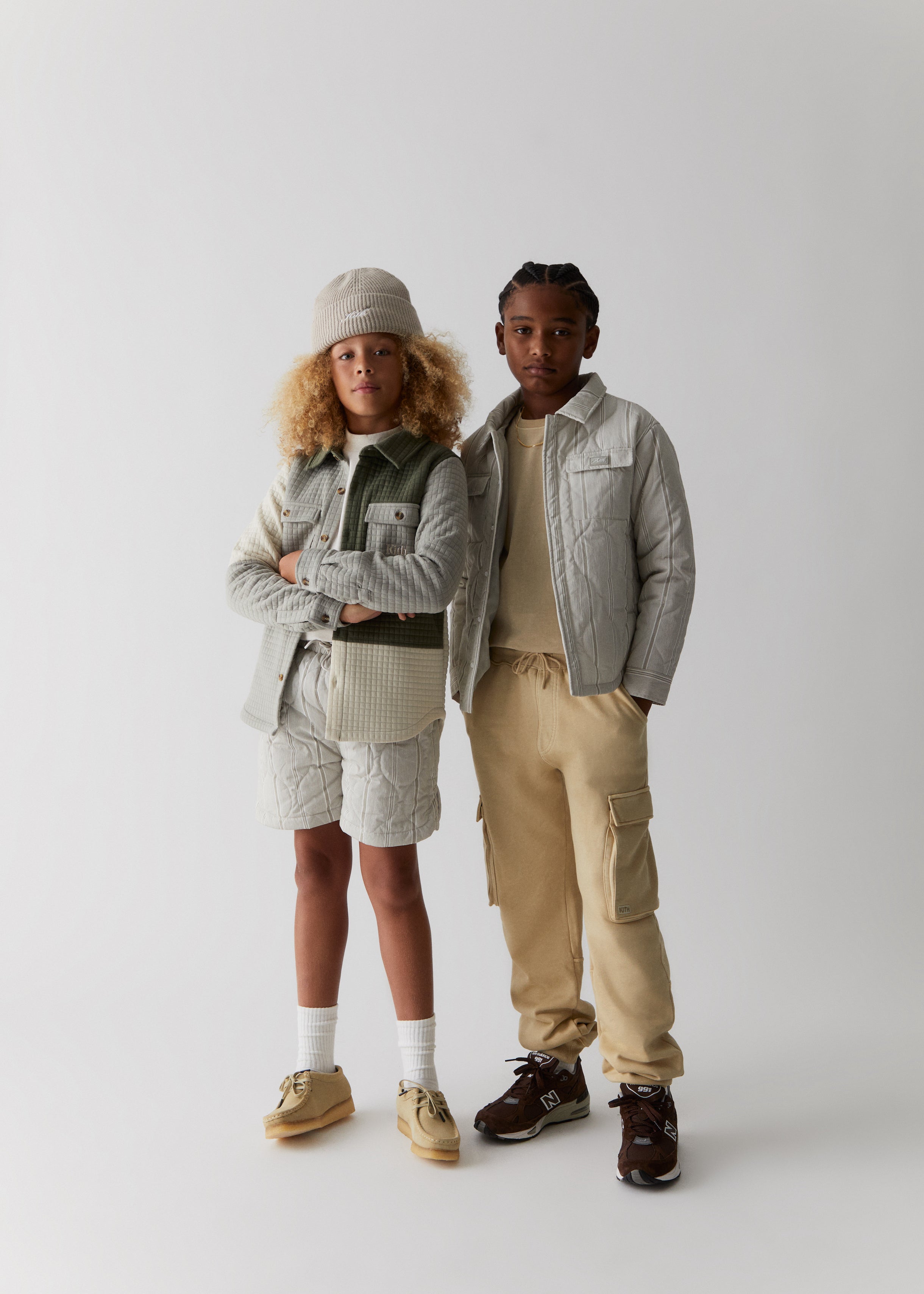 Two kids wearing items from Kith Kids Fall Classics 2023, including a beanie, a corduroy jacket, a quilted shirt jacket, matching quilted shorts, and cargo pants.