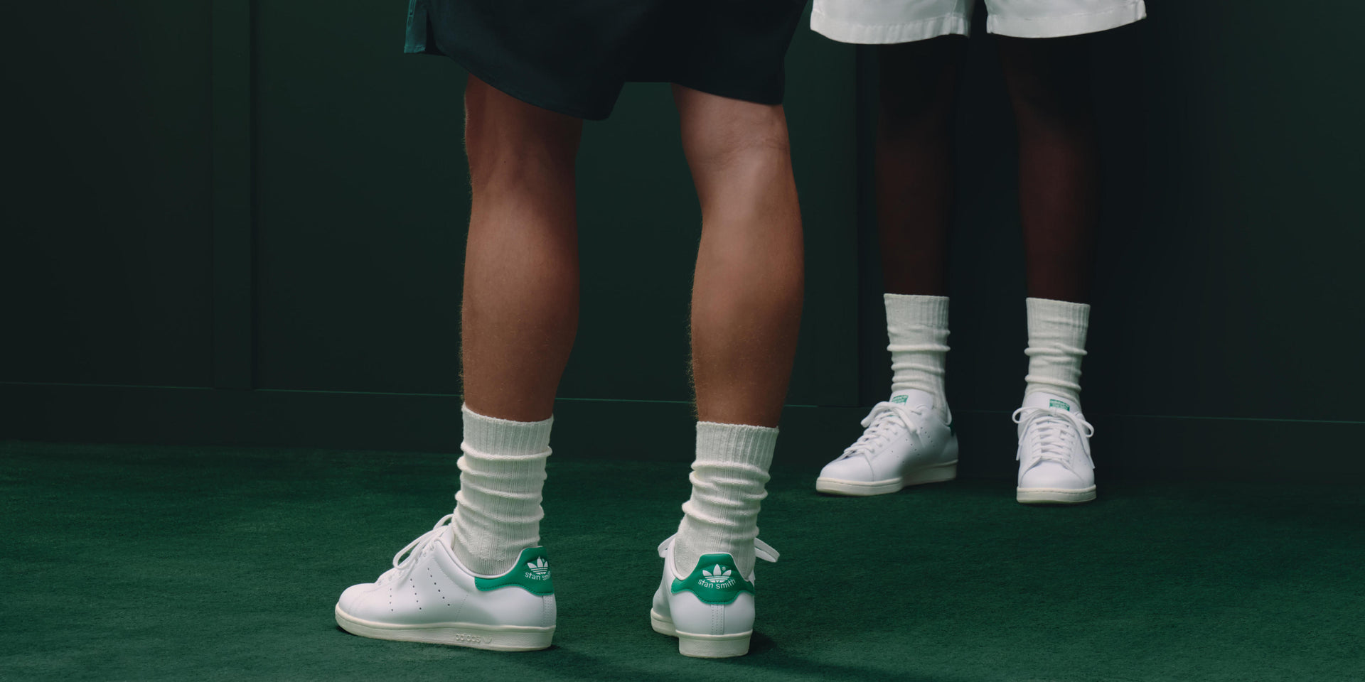 Two people in white Adidas Stan Smith sneakers and slouchy socks stand on a dark green court, legs shown