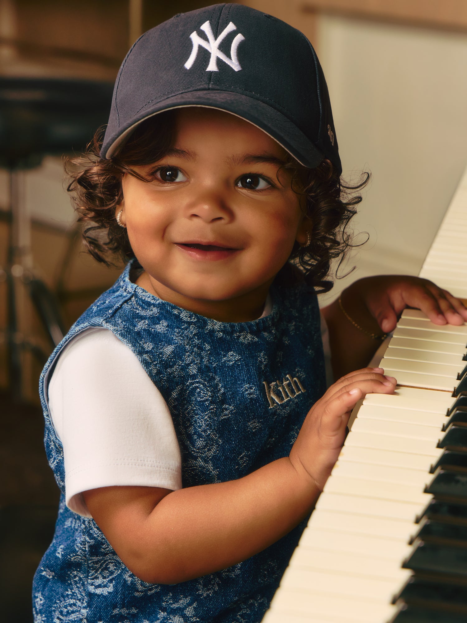 Close-up of a smiling toddler in a navy NY Yankees cap and denim paisley Kith romper over a white tee, hands on grand piano keys