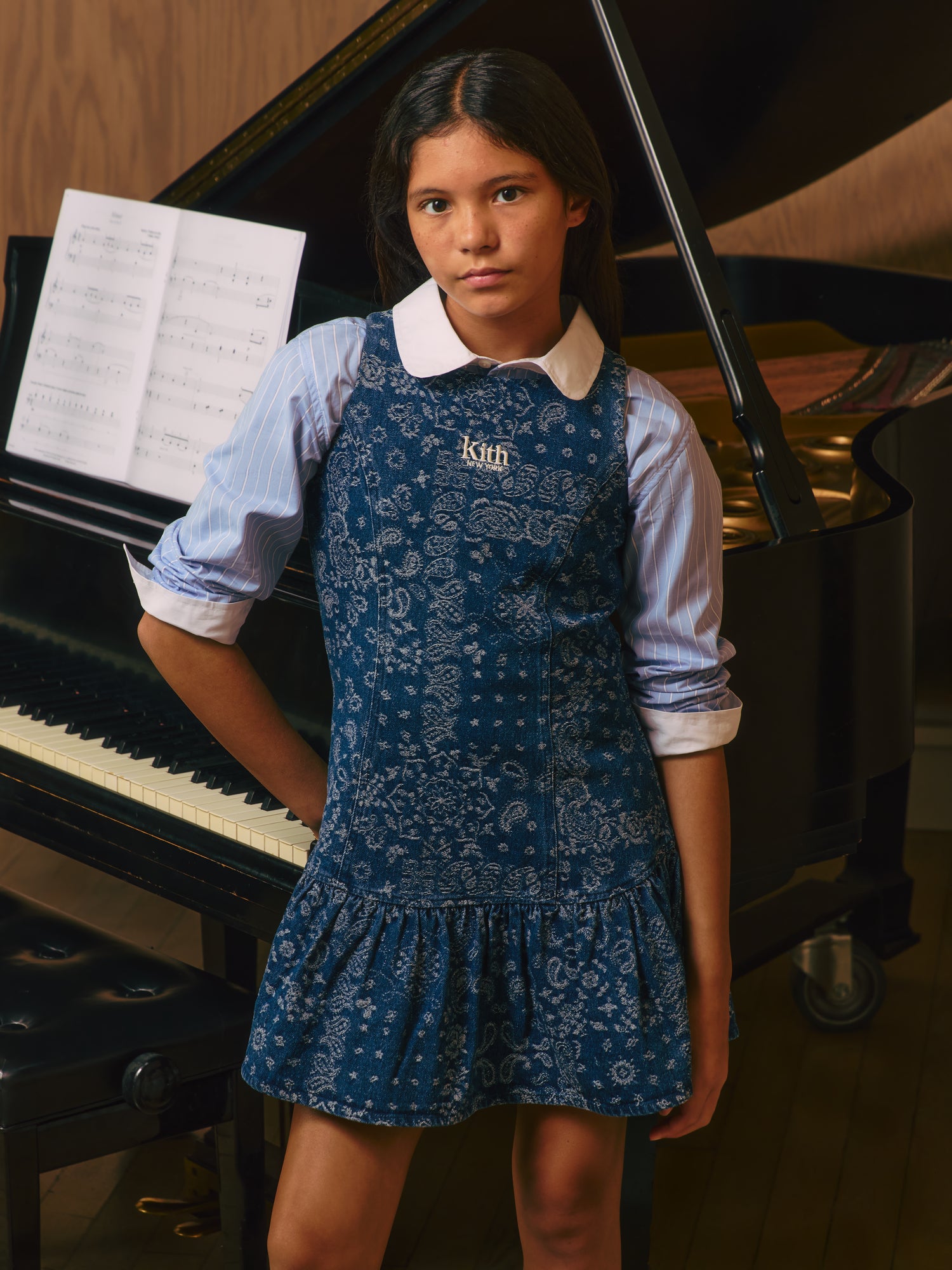 Girl at a grand piano in a denim paisley Kith New York pinafore dress layered over a blue pinstripe shirt with a white collar