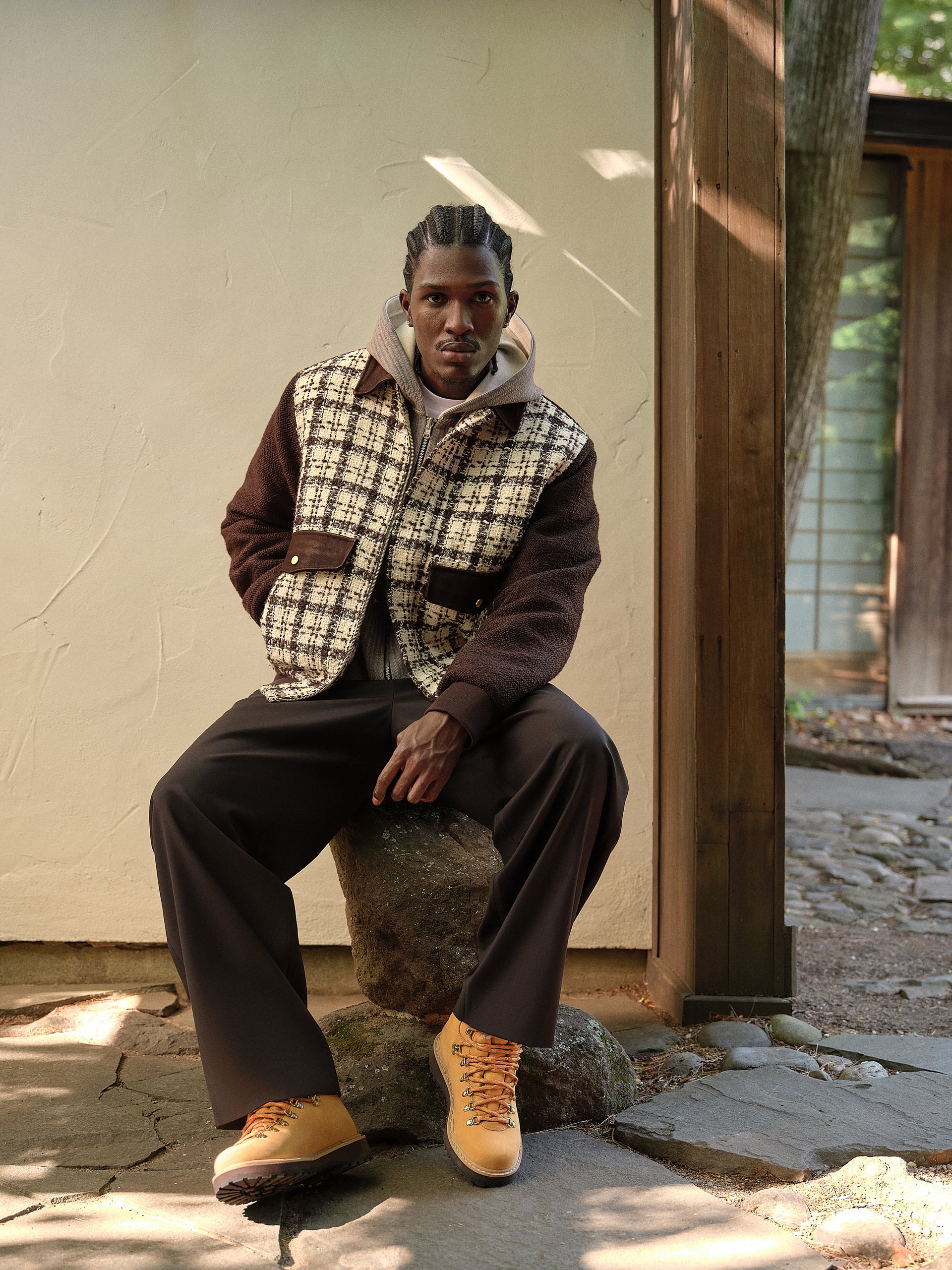 Model in a brown and cream check jacket and dark trousers seated on a rock, wearing tan Kith hiking boots