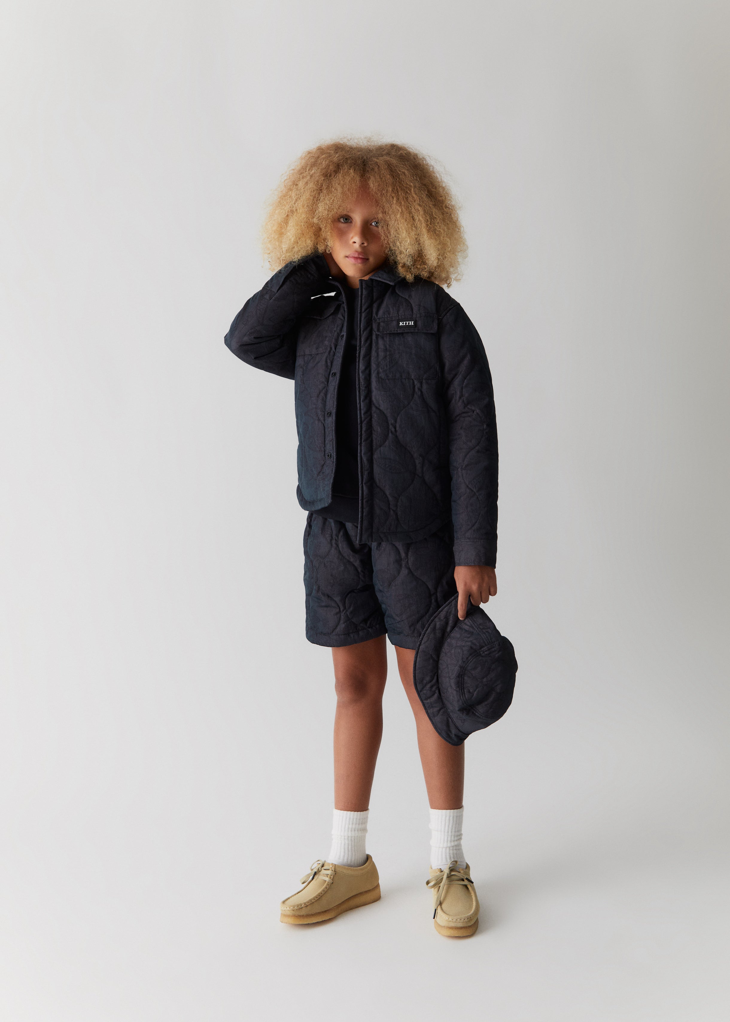 A child wearing a navy quilted shacket with matching navy quilted shorts, holding a navy quilted bucket hat from the Kith Kids Fall Classics 2023 collection.