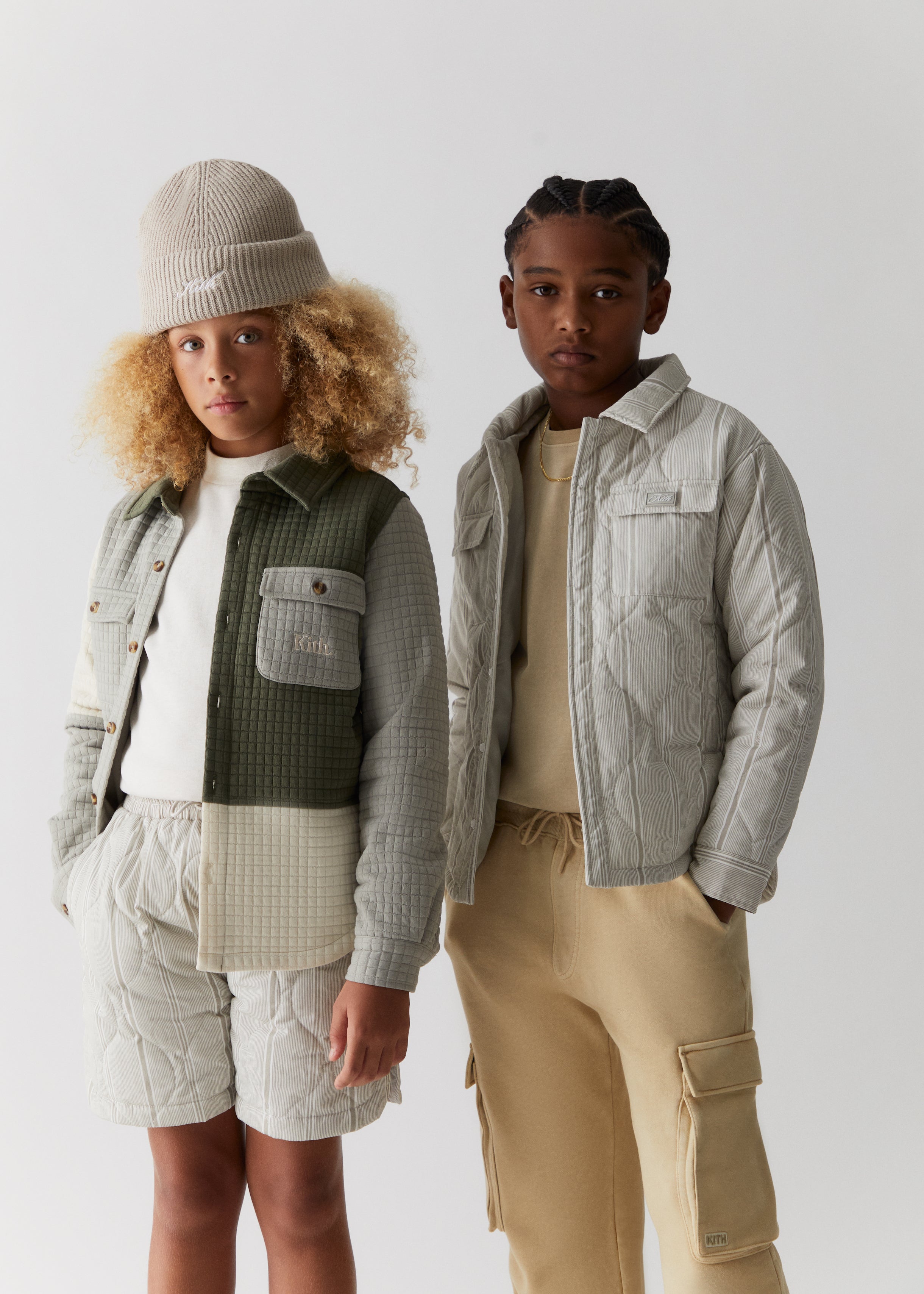 Two kids wearing clothing from the Kith Kids Fall Classics 2023 collection, including a grey knit Kith script beanie, a color-blocked quilted shirt jacket, grey quilted shorts, a grey quilted shacket, and tan cargo pants. 