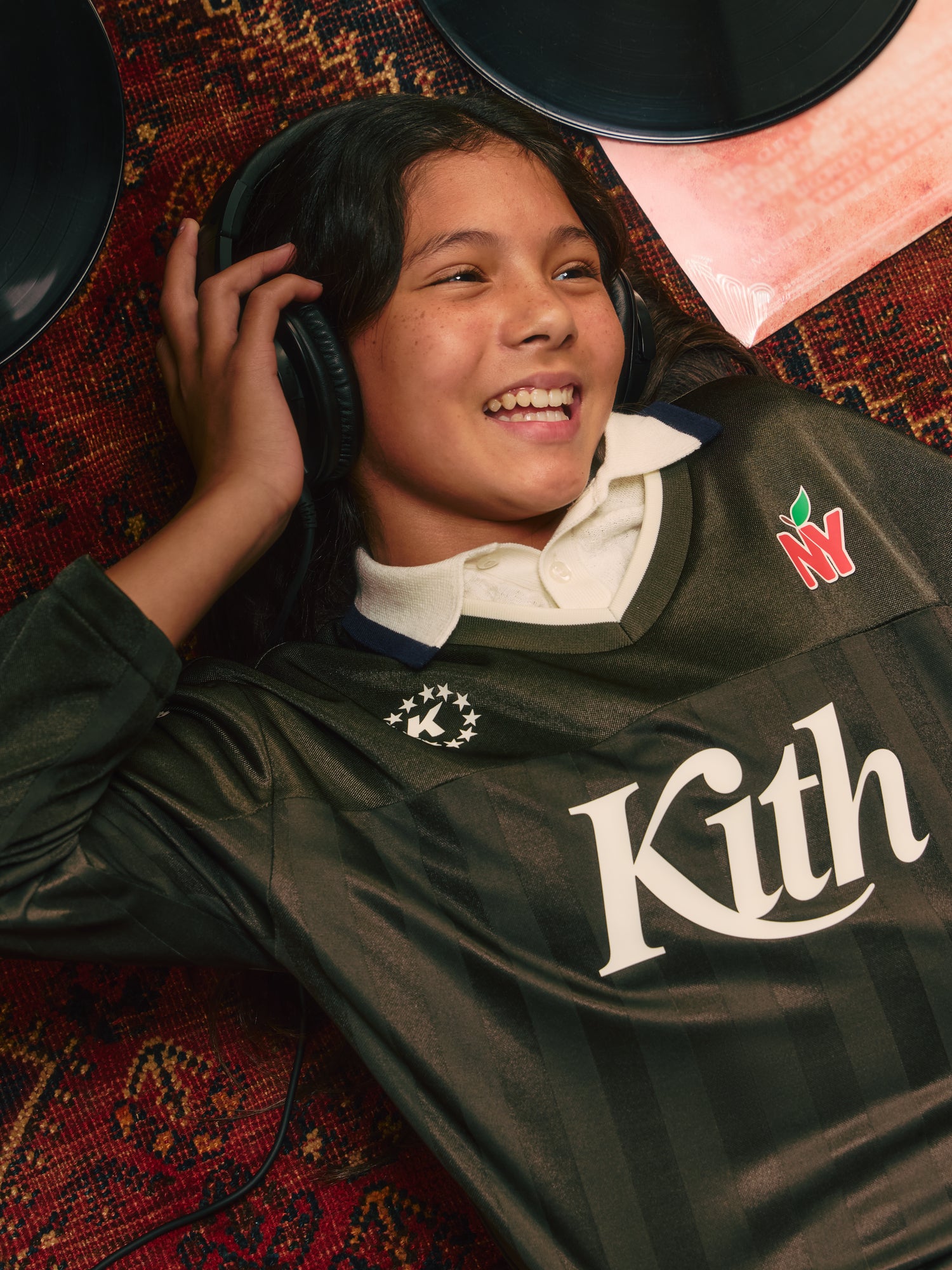 Smiling girl in black headphones and olive Kith NY soccer jersey lying on a red patterned rug beside vinyl records