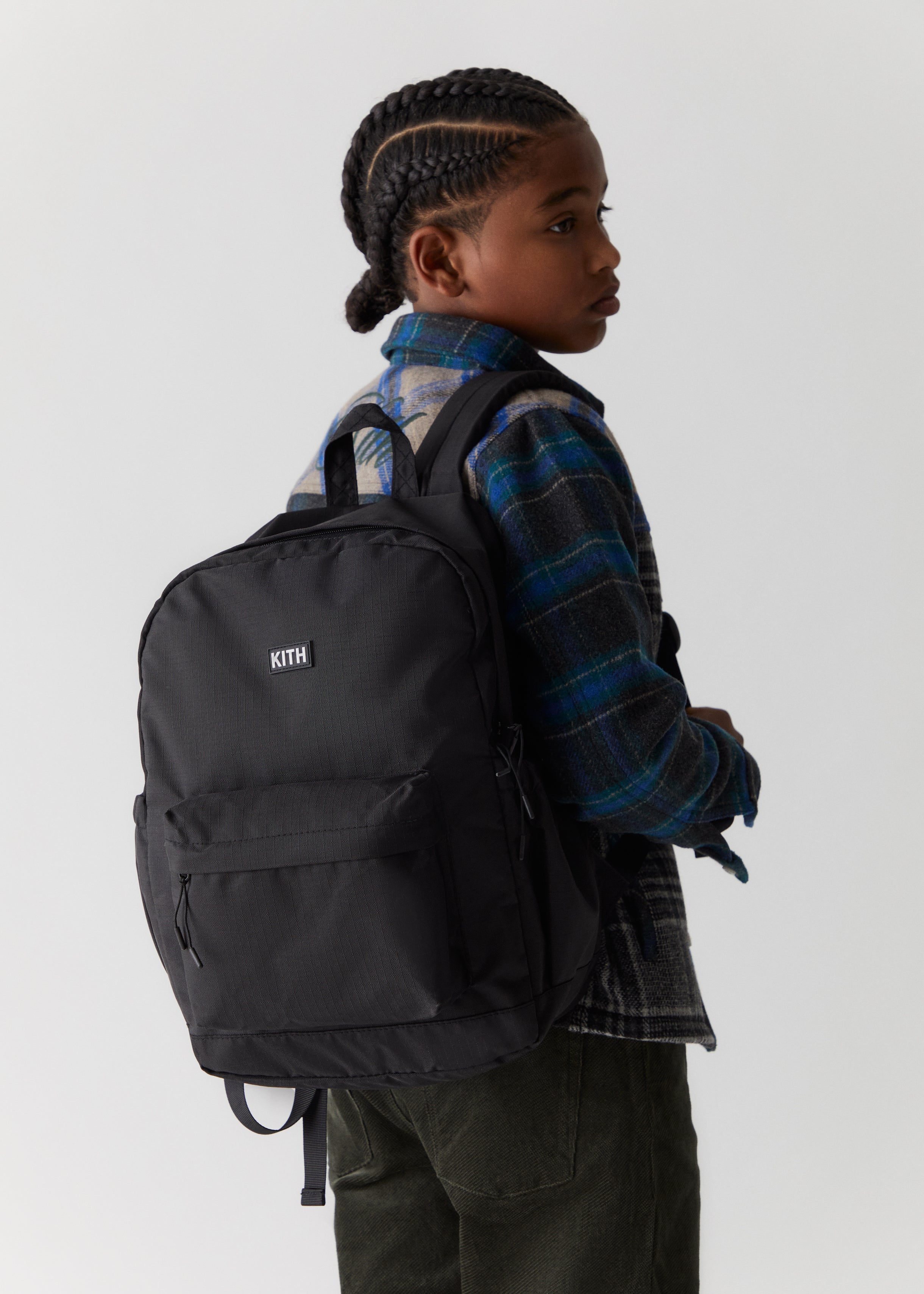 A child wearing a black Kith branded backpack and blue flannel shirt from the Kith Kids Fall Classics 2023 collection.