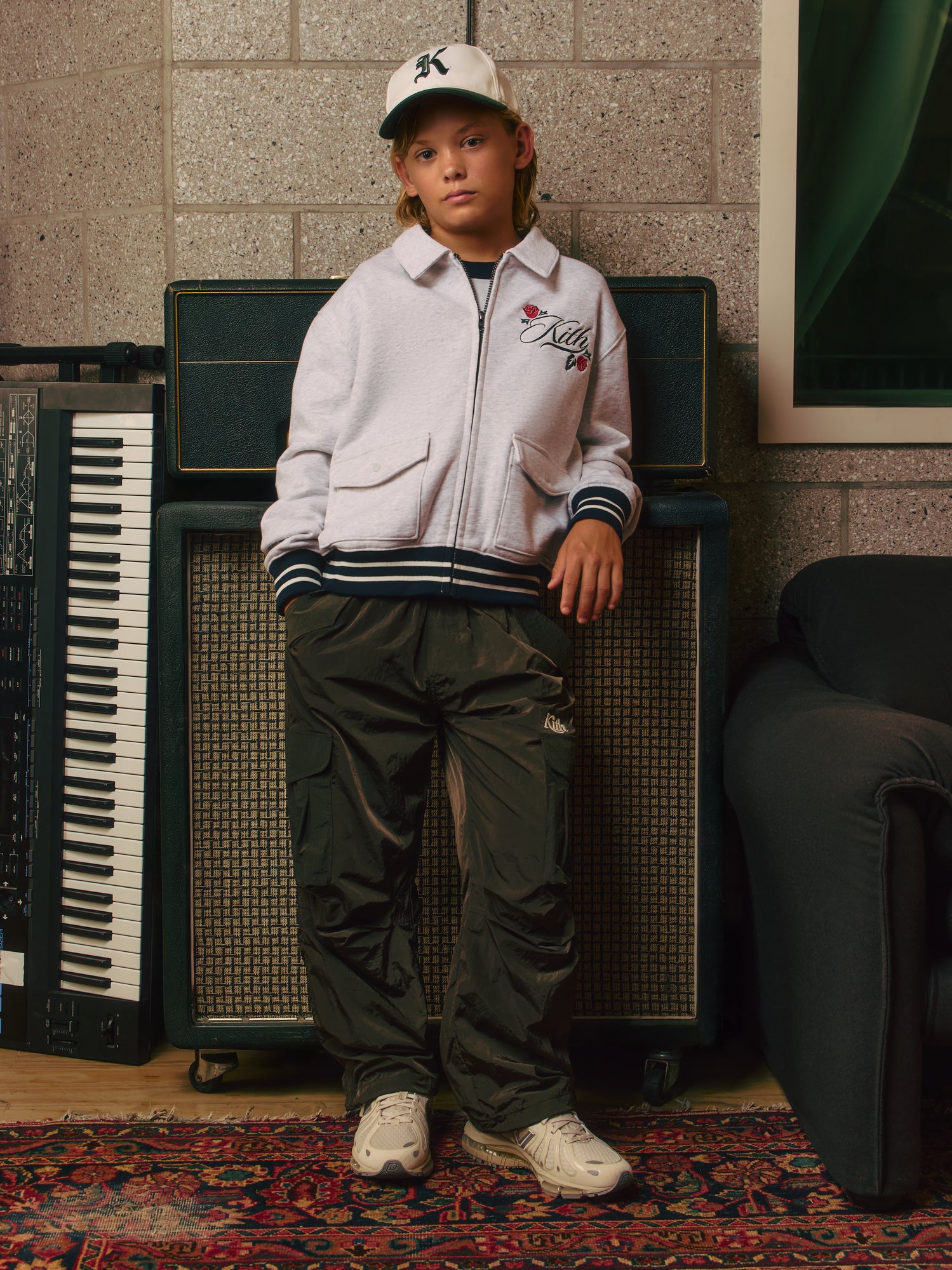 Blond boy in a white K-logo Kith cap and grey rose-embroidered zip sweatshirt with dark green cargo pants and white sneakers, amps behind