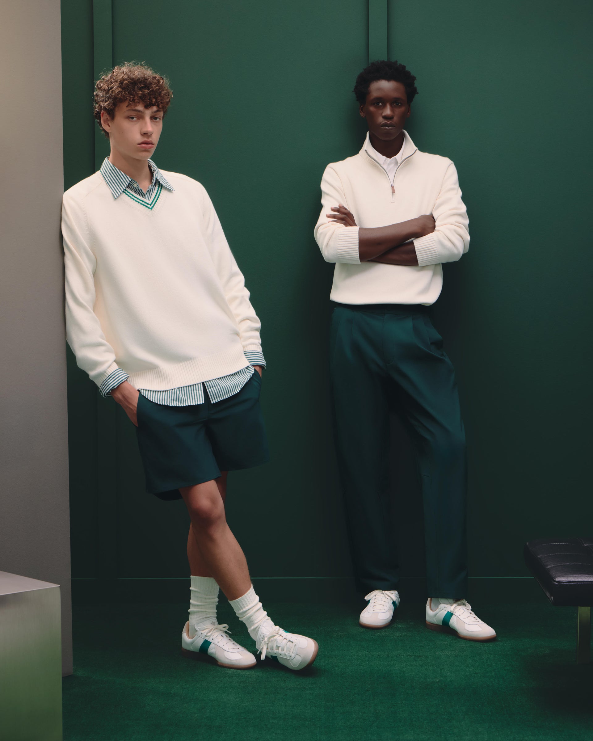 Two male models in cream sweaters with green shorts and trousers and white sneakers against a deep green wall