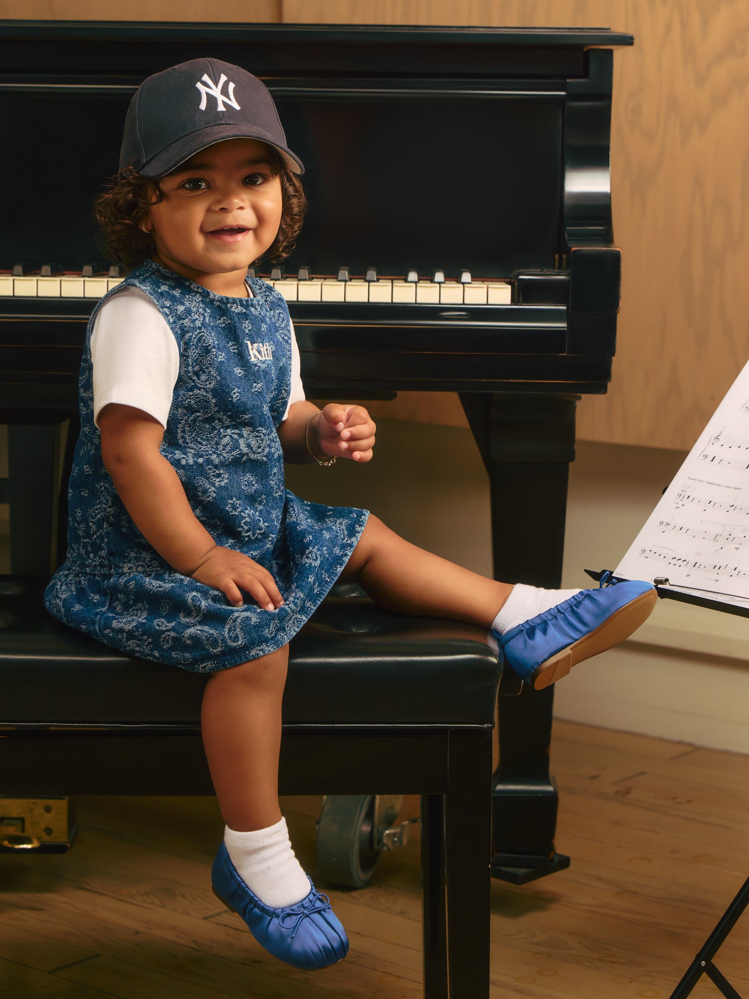 Smiling toddler on a piano bench in a navy Yankees cap and denim paisley Kith pinafore over a white tee, blue satin flats, sheet music
