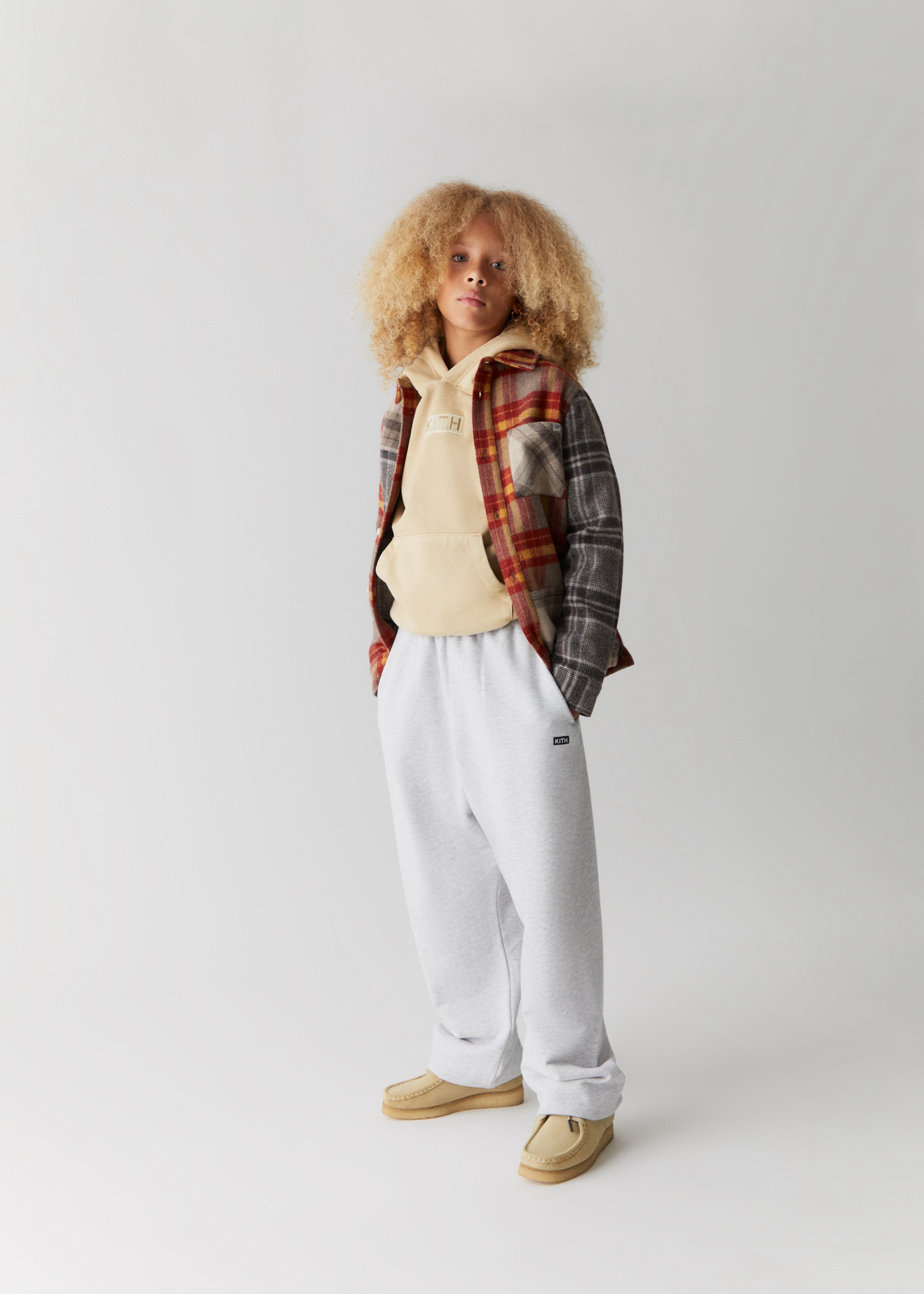 A child wearing a color-blocked grey and red flannel shirt over a yellow Kith hoodie and grey sweatpants, all from the Kith Kids Fall Classics 2023 collection.