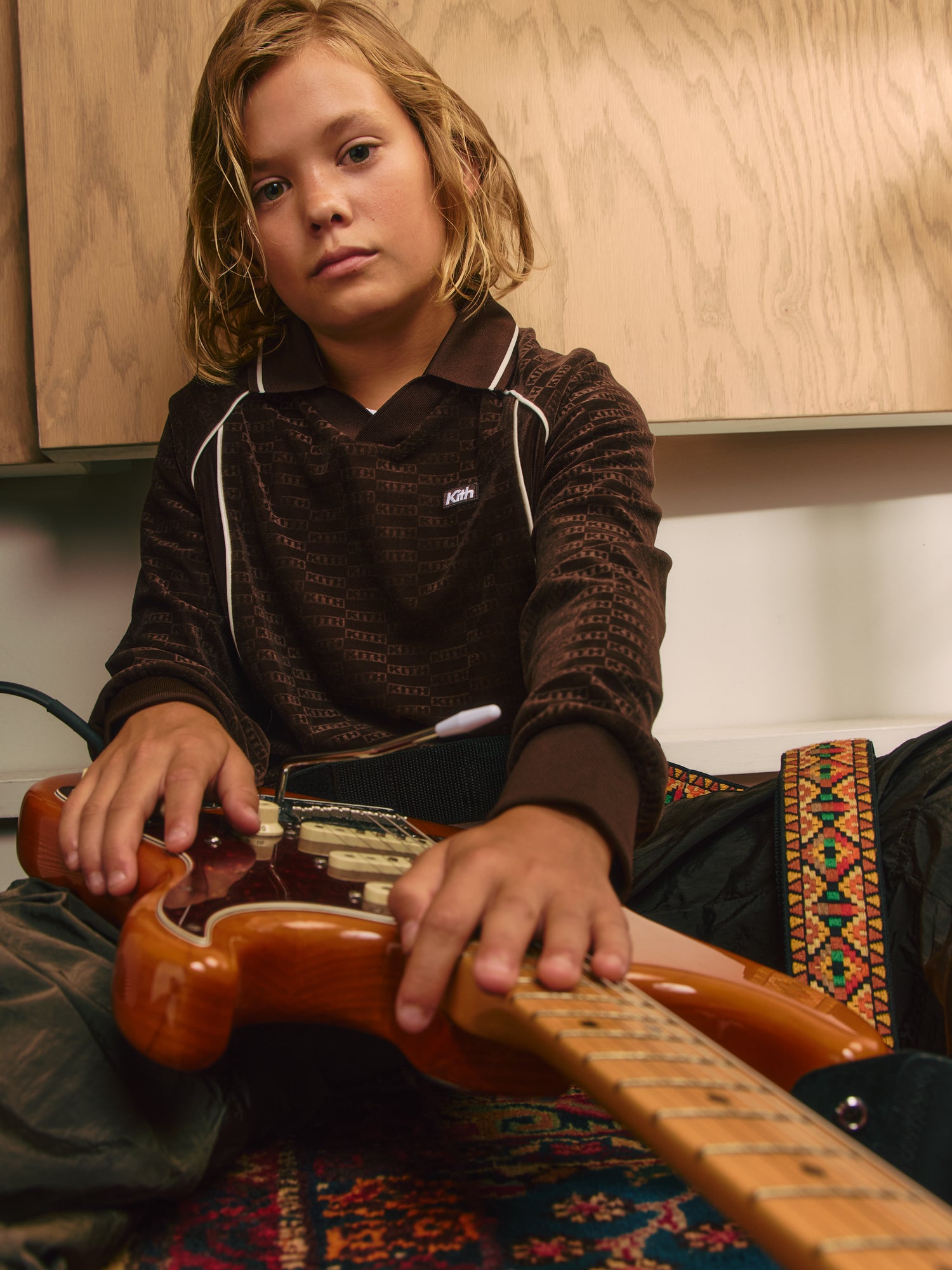 Low-angle of a blond boy on a patterned rug holding a natural-wood electric guitar, wearing a brown Kith monogram long-sleeve polo