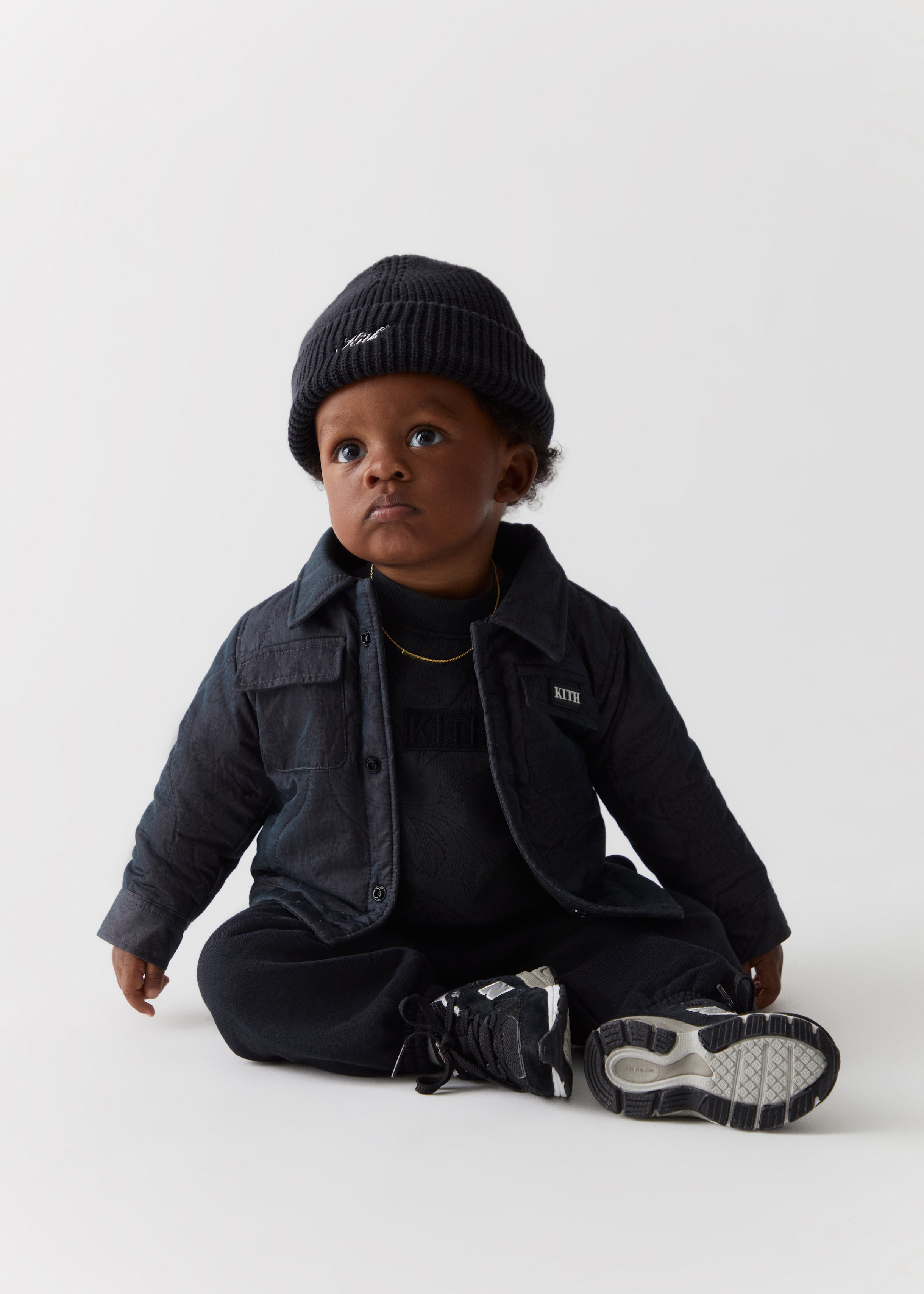 A baby wearing a black Kith script beanie, with a black Kith-branded shirt jacket from the Kith Kids Fall Classics 2023 collection.