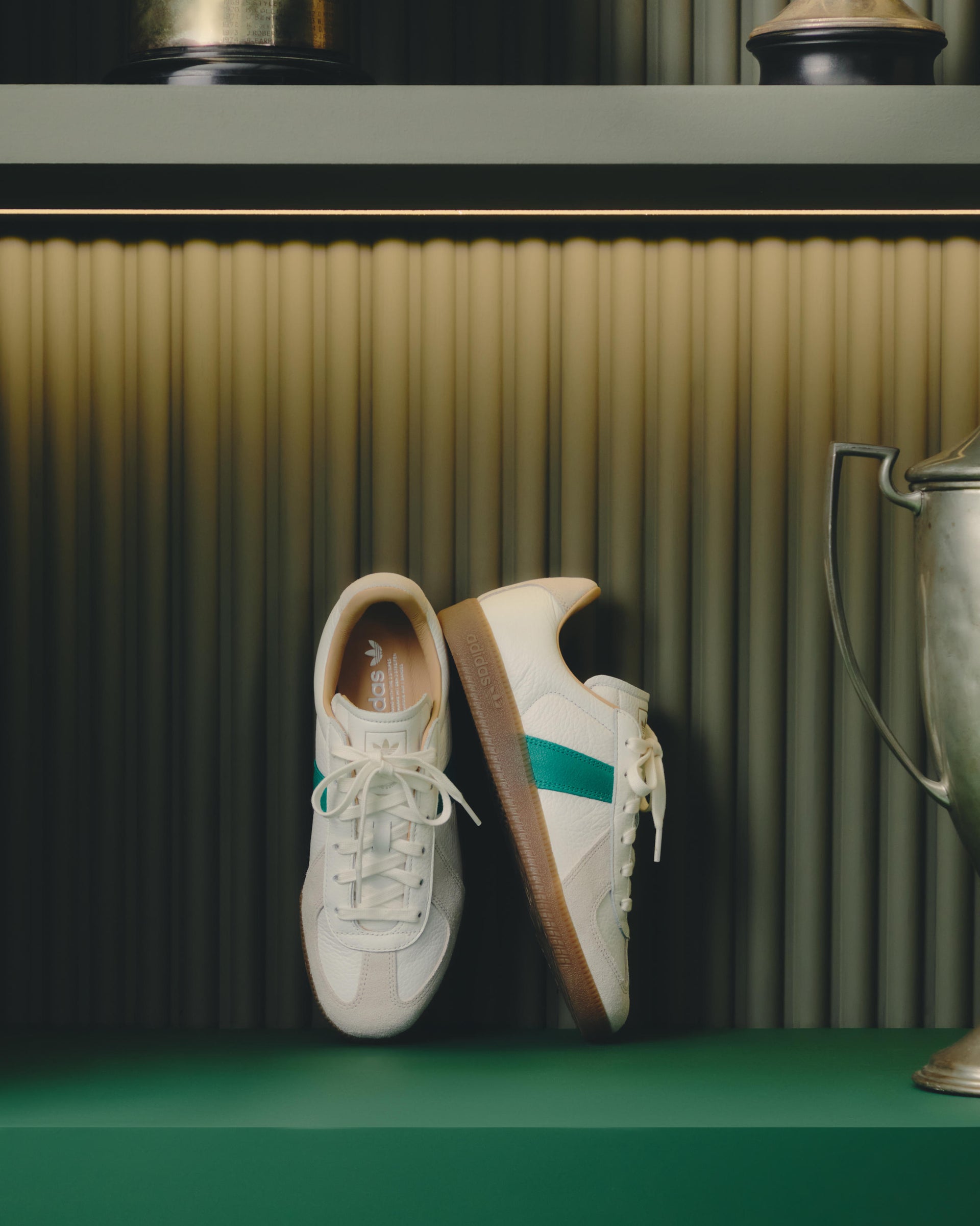 Pair of cream leather Adidas sneakers with green stripe and gum sole displayed on a green shelf beside a silver trophy