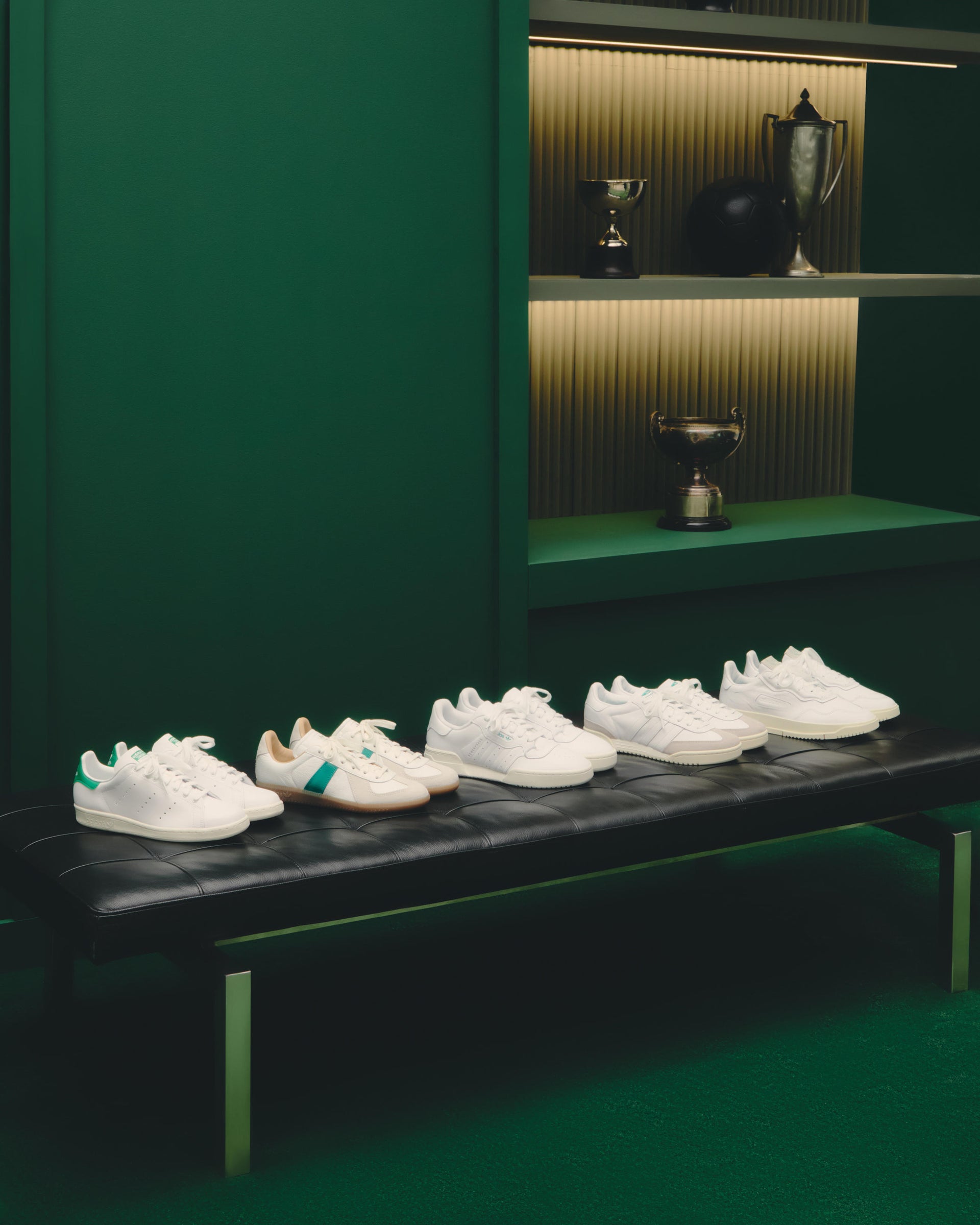 Five white leather sneakers lined up on a black leather bench in a dark green trophy room, angled side view