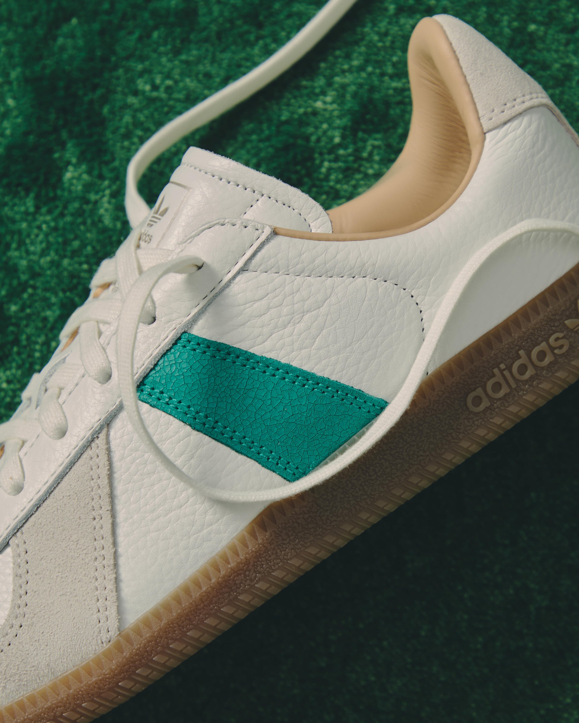 Close-up of a cream leather Adidas sneaker with green stripe, gold branding and gum sole on green turf