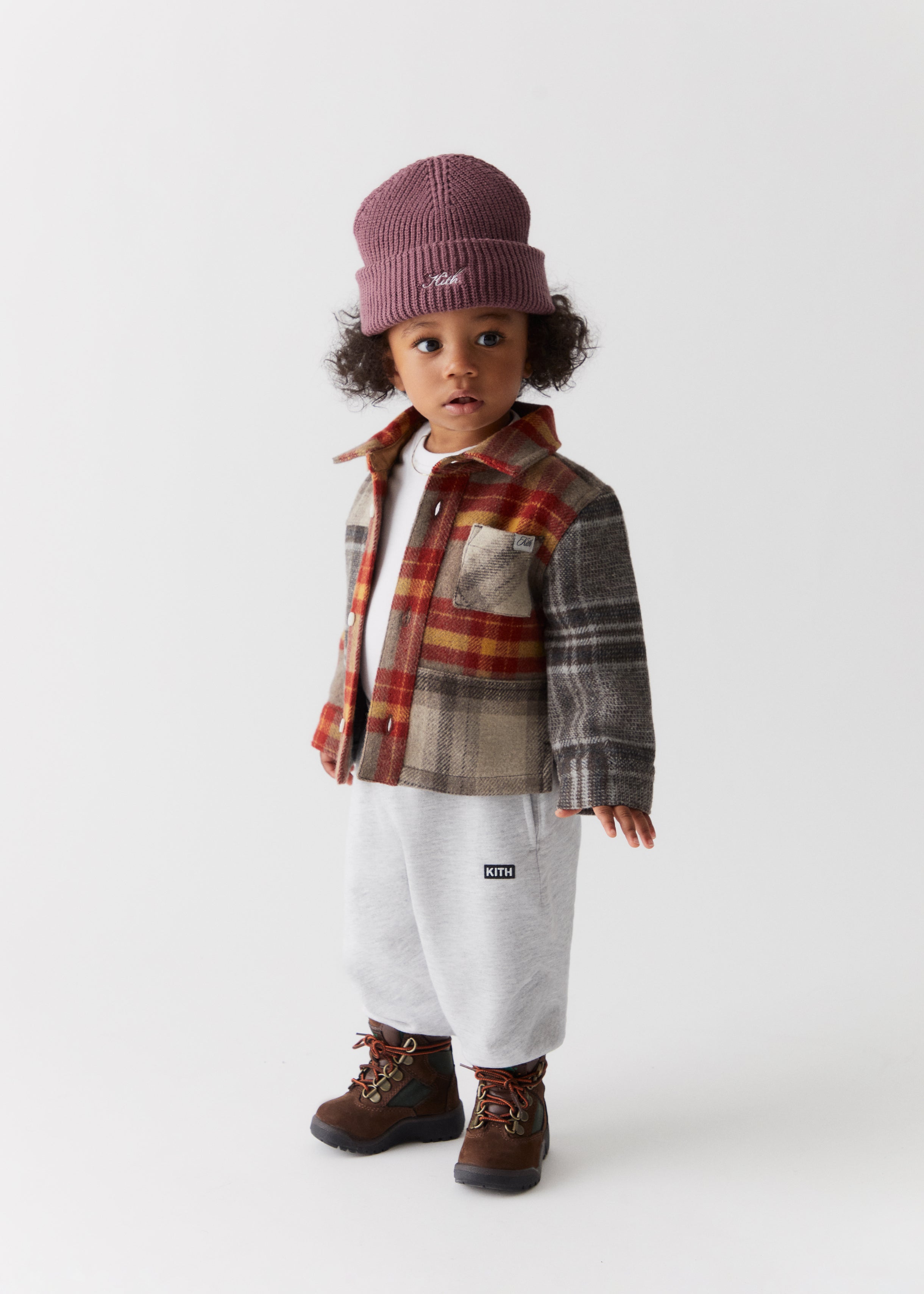 A toddler wearing items from Kith Kids Fall Classics 2023, including a knit logo beanie, plaid shirt jacket, and grey sweatpants.