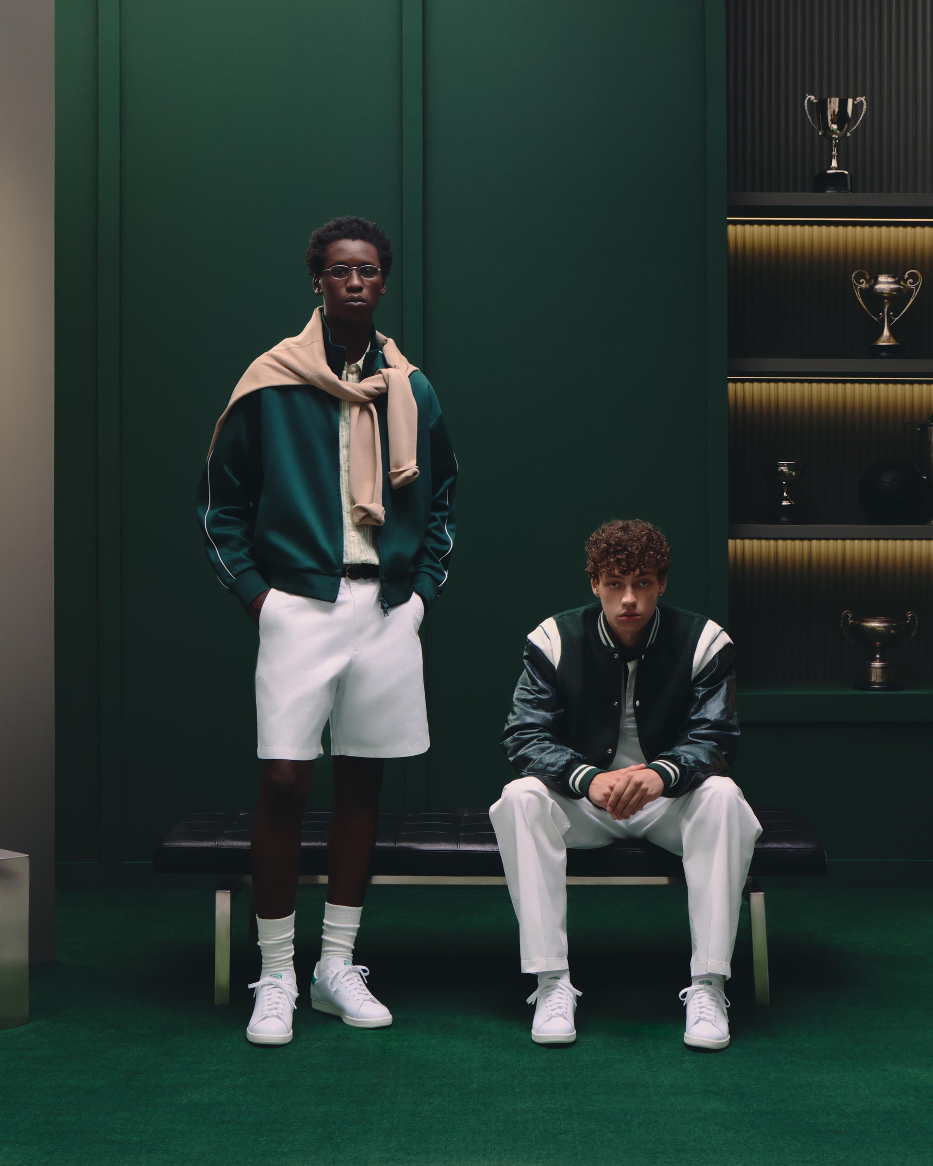 Two men in a green track jacket and a varsity jacket with white shorts and pants in a dark green trophy room