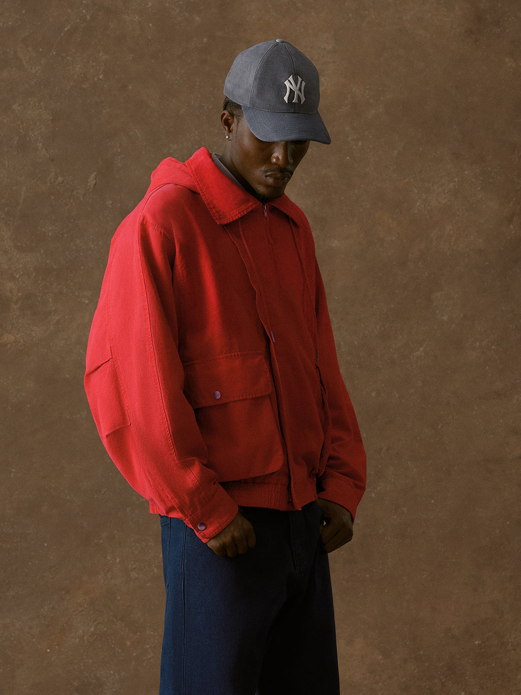 Model wearing red hooded harrington jacket, grey NY Yankees cap, and dark denim jeans against brown backdrop