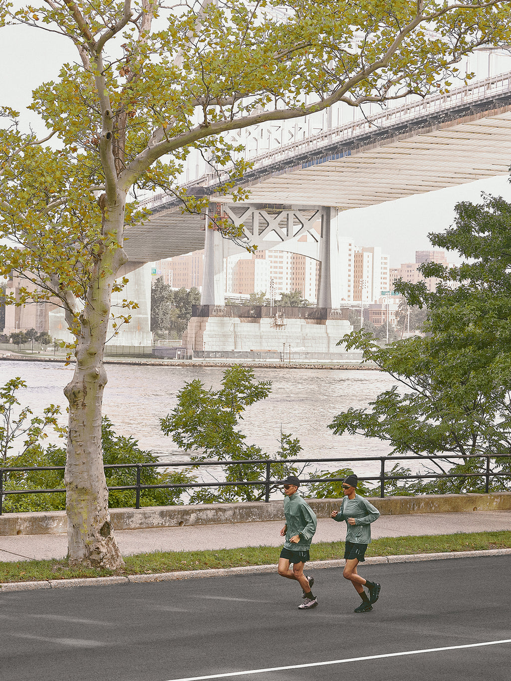 Two Kith Training runners in sage green pullovers and black shorts jogging on a riverside road beneath a large bridge, city skyline behind