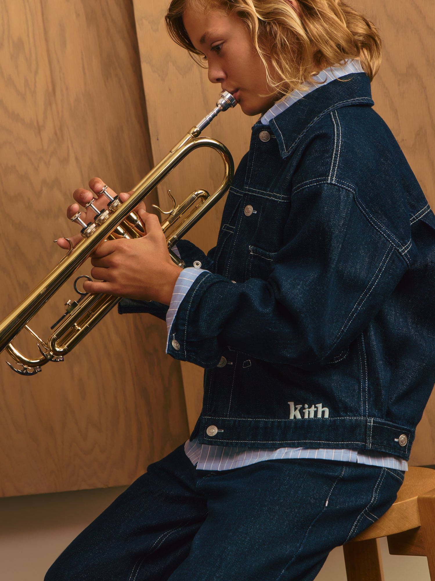 Blond boy in dark indigo Kith denim jacket and jeans over a pinstripe shirt playing a brass trumpet against wood paneling