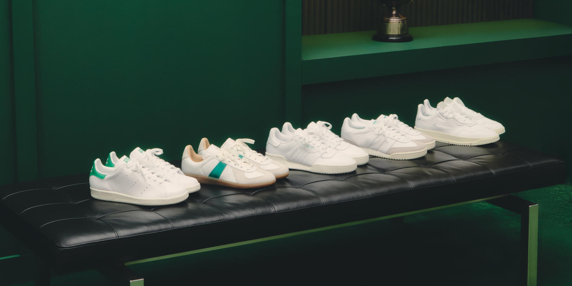 Five pairs of white leather sneakers from the adidas x Kith collection lined up on a black leather bench in a dark green room
