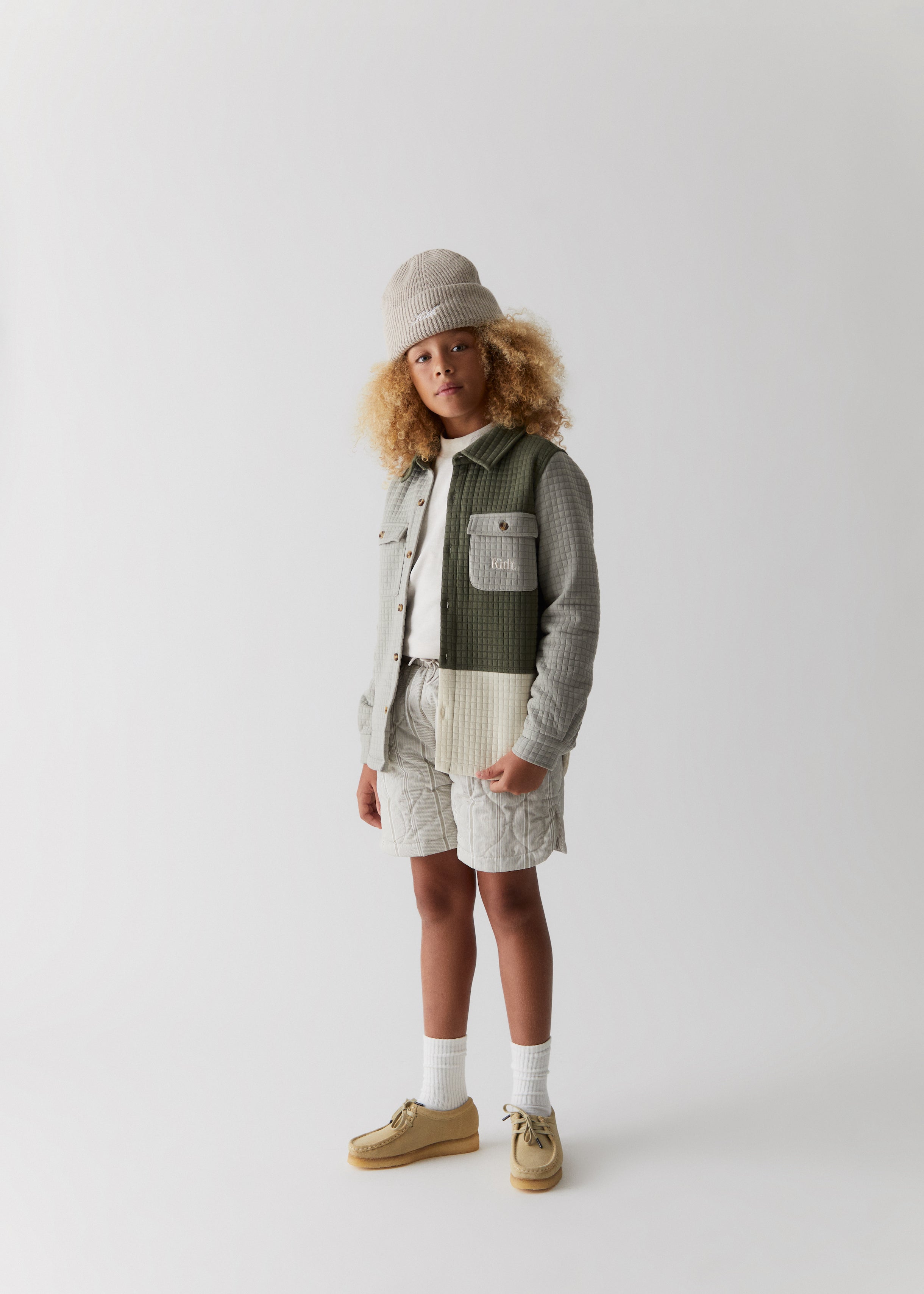A child wearing a color-blocked corduroy shirt jacket, grey quilted shorts, and a grey Kith script knit beanie from the Kith Kids Fall Classics 2023 collection.