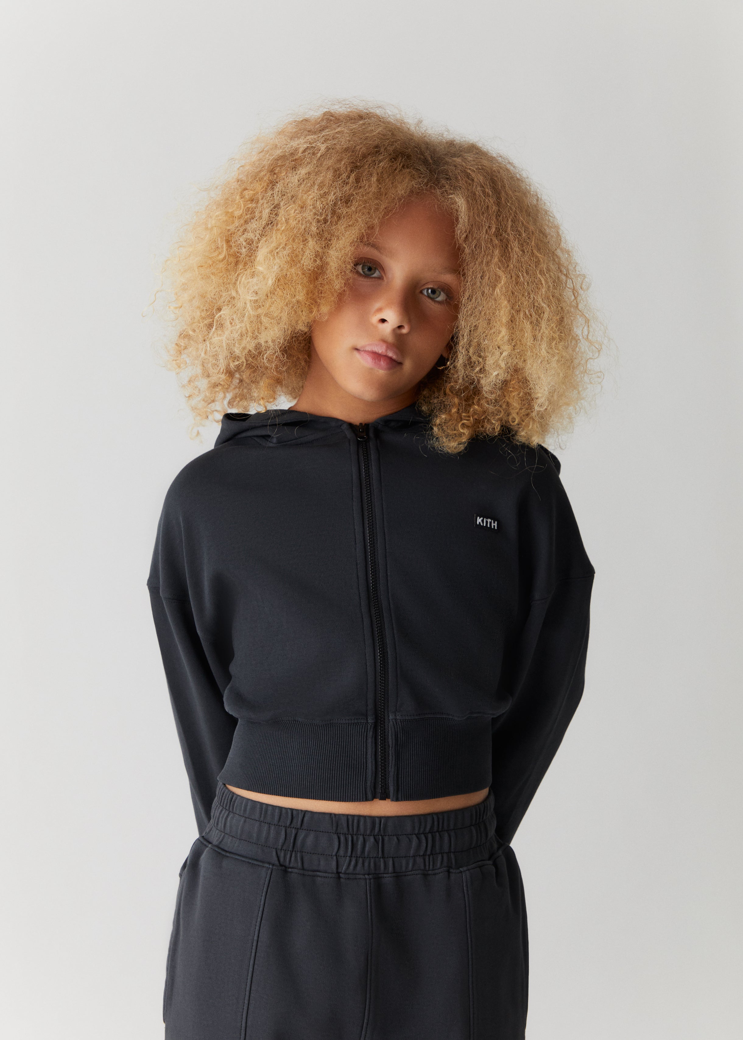 A child wearing a black zip-up hoodie and black sweatpants from the Kith Kids Fall Classics 2023 collection.