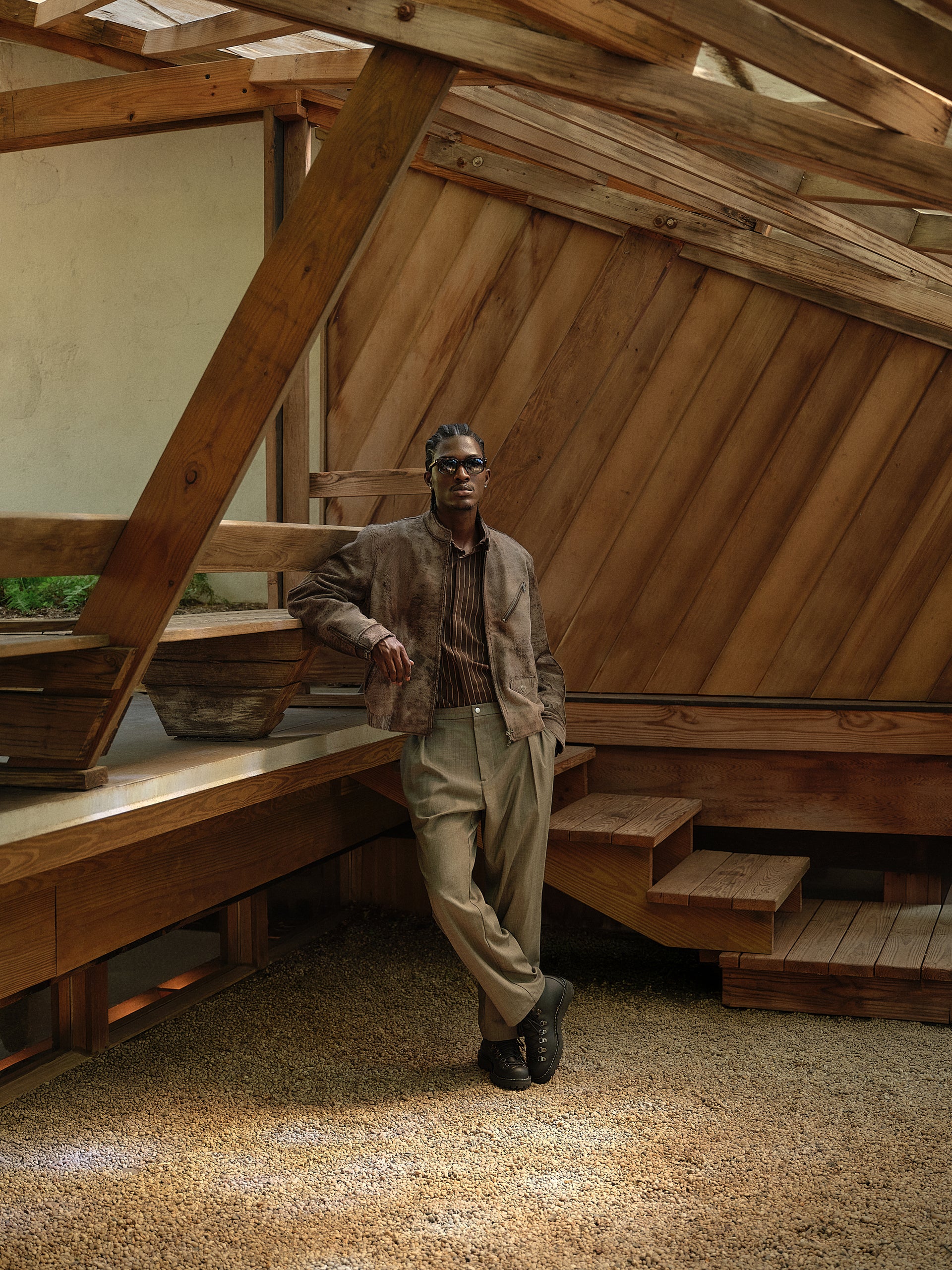 Model in a weathered brown suede jacket, striped shirt and taupe trousers leaning against angular wood architecture