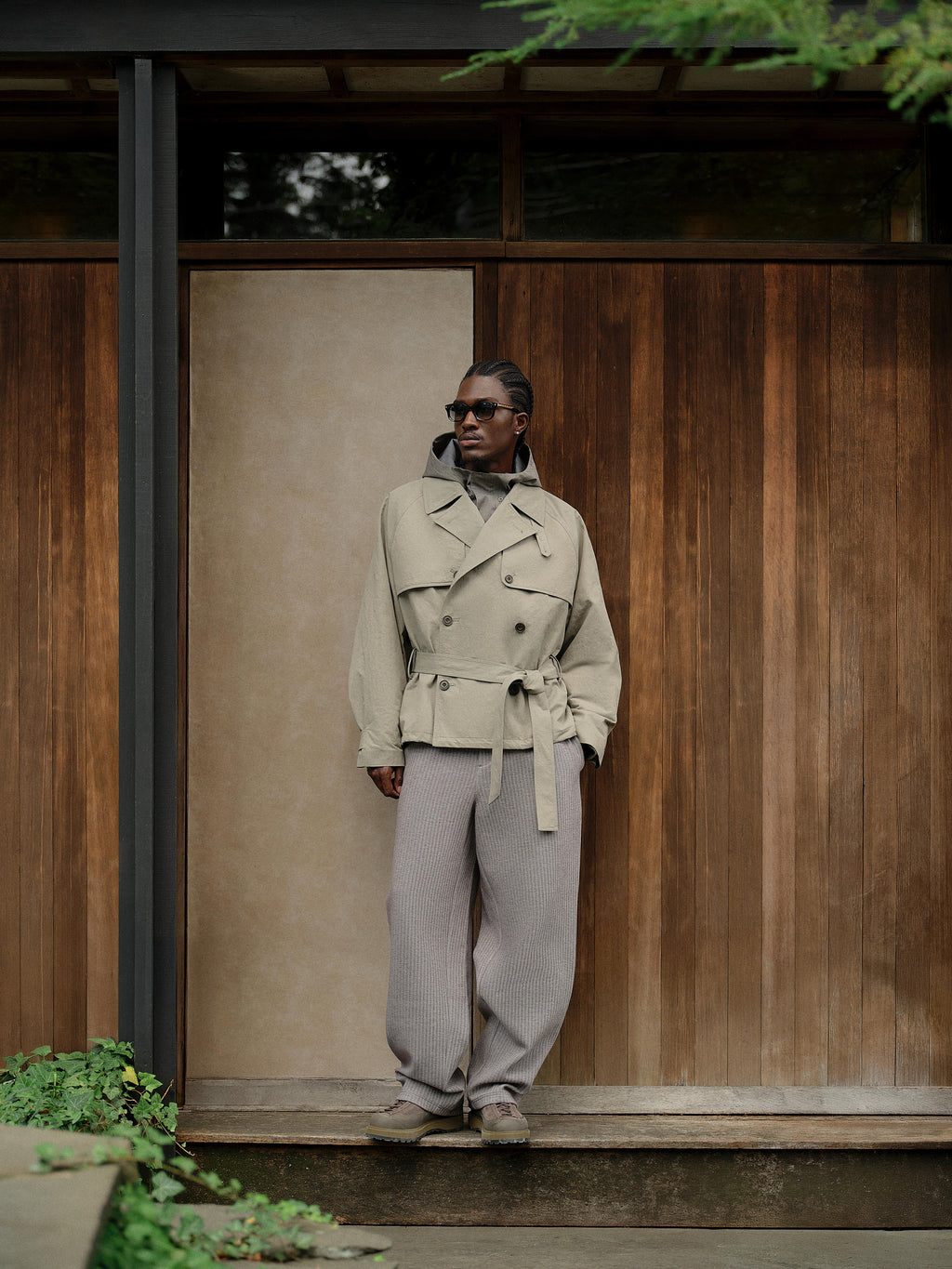 Man in sunglasses at a wood-panel wall in a khaki Kith belted jacket, grey hoodie and ribbed wide-leg trousers, full-length vertical shot