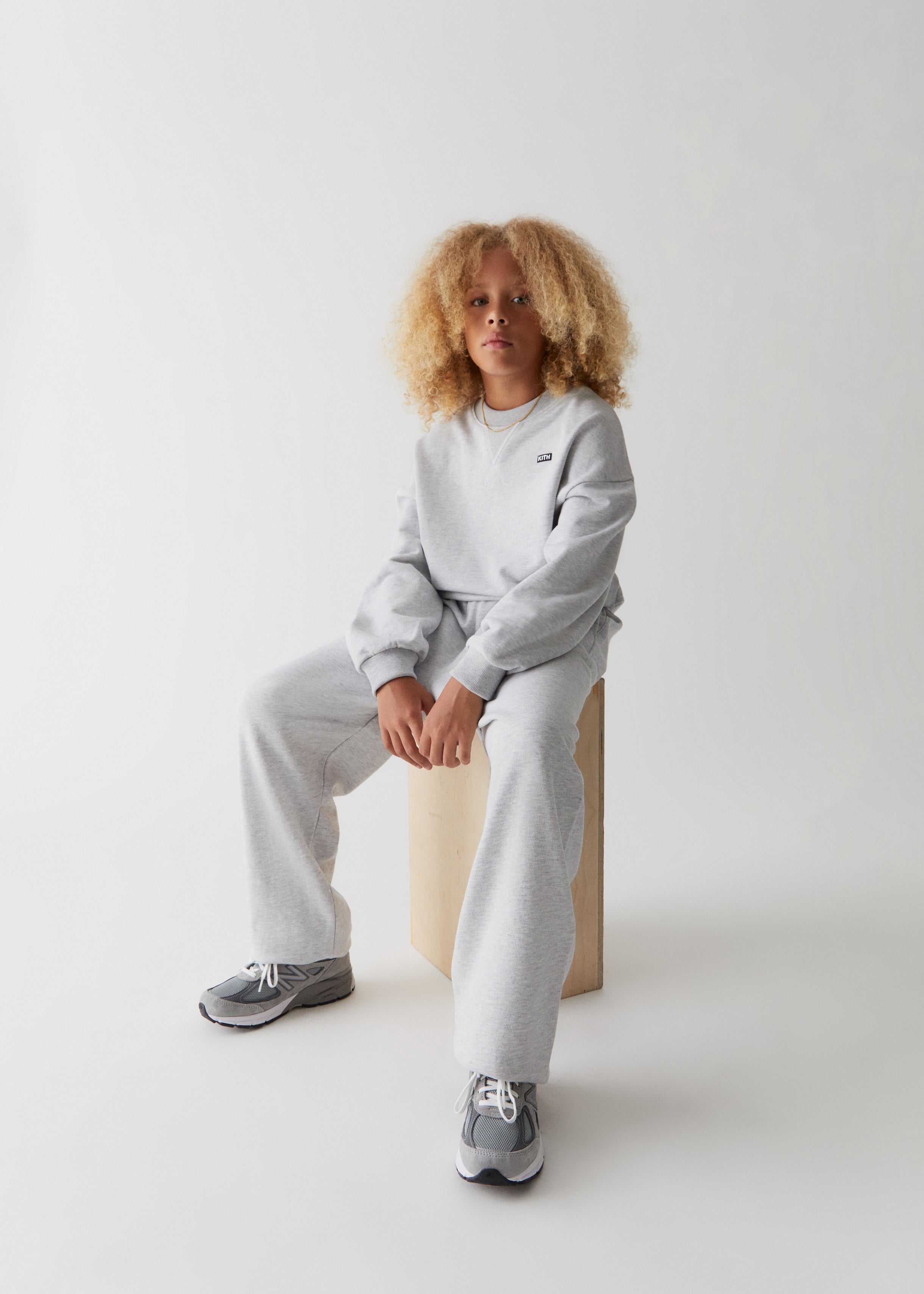 A child sitting while wearing a matching grey Kith logo crewneck and sweatpants. 
