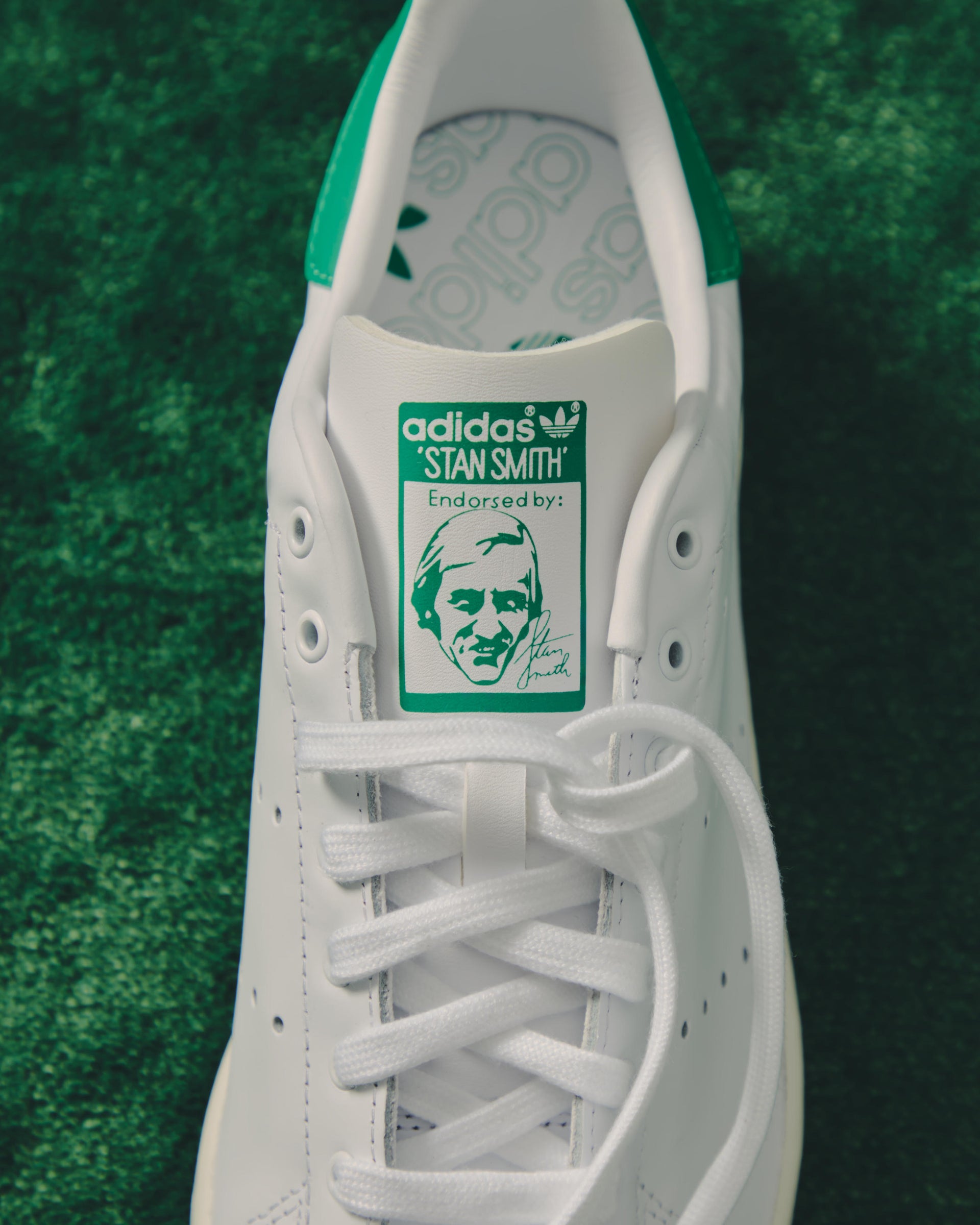 Close-up of adidas Stan Smith white leather sneaker tongue with green Stan Smith portrait label, on green turf