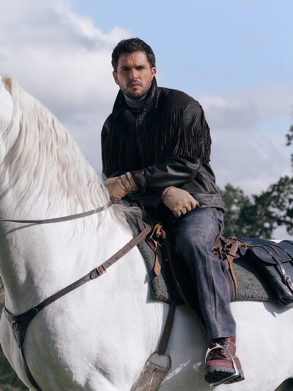 Man seated on a white horse in a black fringed leather Kith jacket, dark jeans and brown boots; Men's Winter campaign, vertical crop