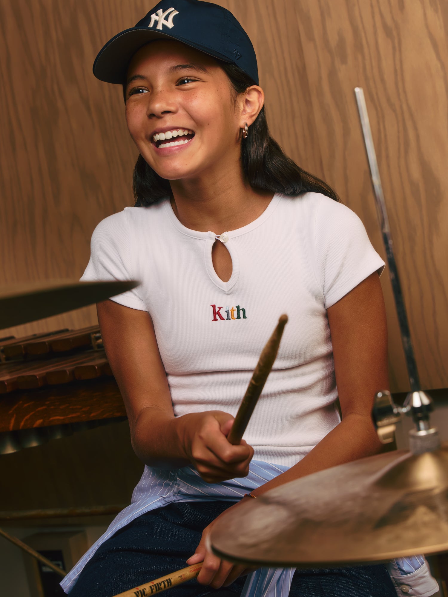 Smiling girl in a navy Yankees '47 cap and white ribbed keyhole Kith tee playing cymbals with drumsticks in a wood-paneled studio